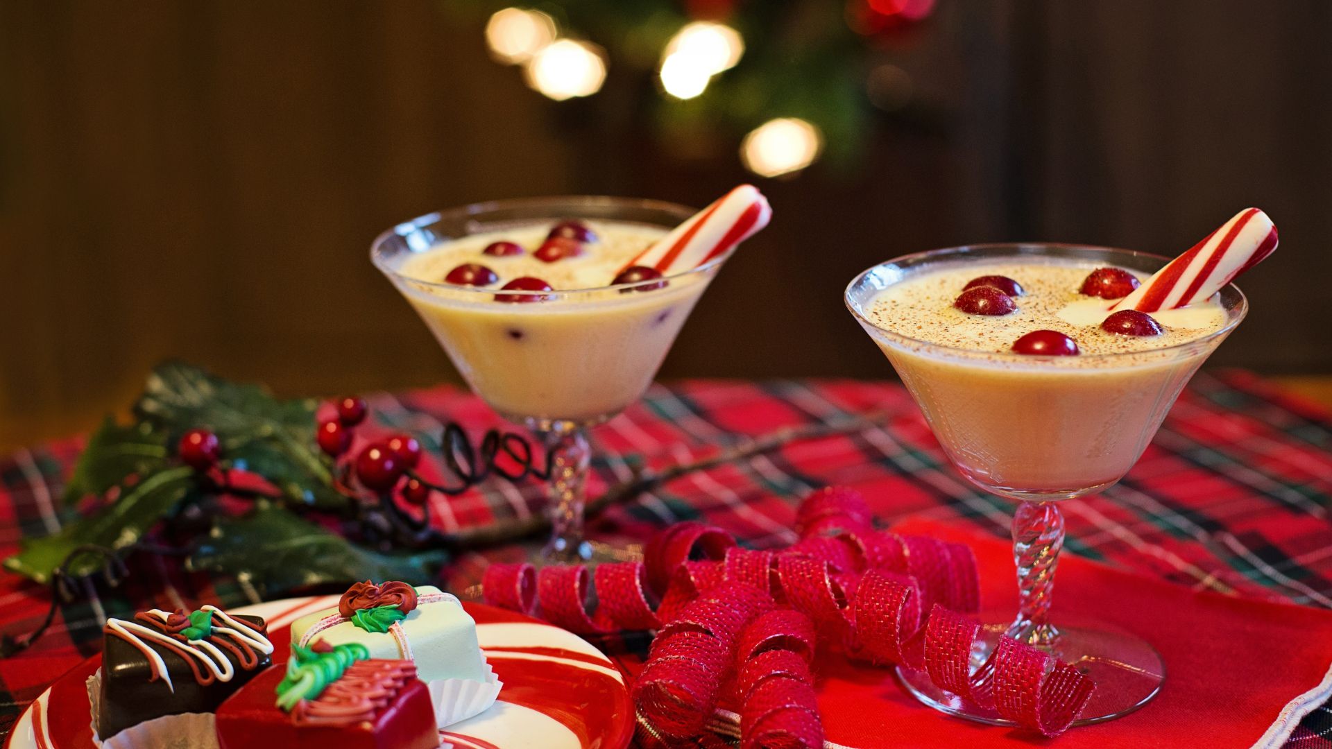 A festive holiday scene featuring two glasses of eggnog garnished with candy canes and cranberries, surrounded by small pastries and red ribbons on a plaid tablecloth, with a Christmas tree in the background.