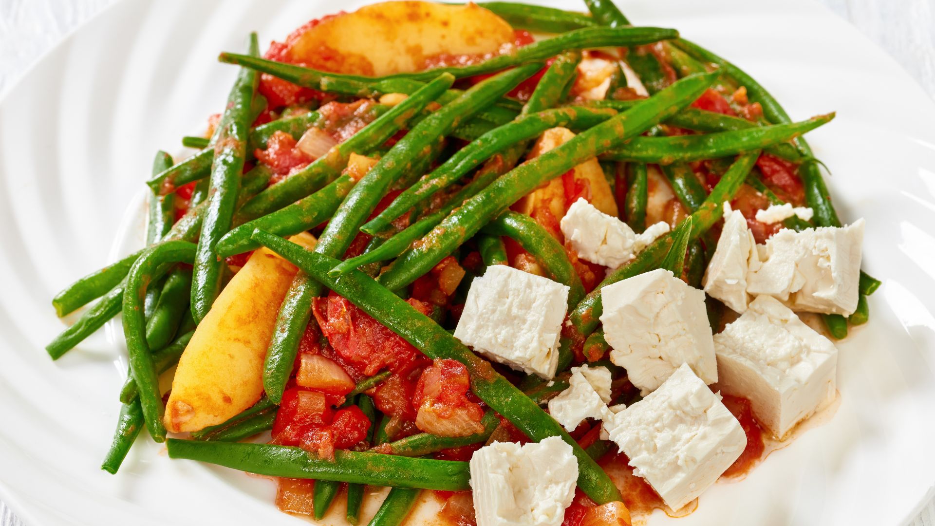 A plate of Fasolakia Lathera, a Greek dish with green beans, tomatoes, and potatoes stewed in olive oil and herbs, topped with crumbled feta cheese.