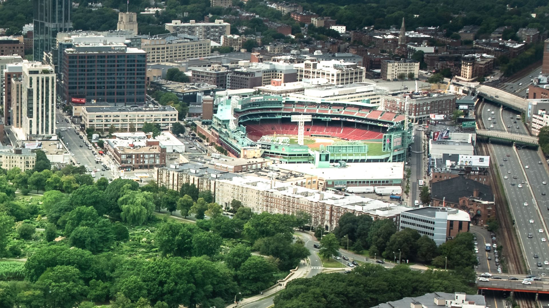 An aerial view of Fenway Park, a baseball stadium in Boston, Massachusetts, surrounded by city buildings and a highway.