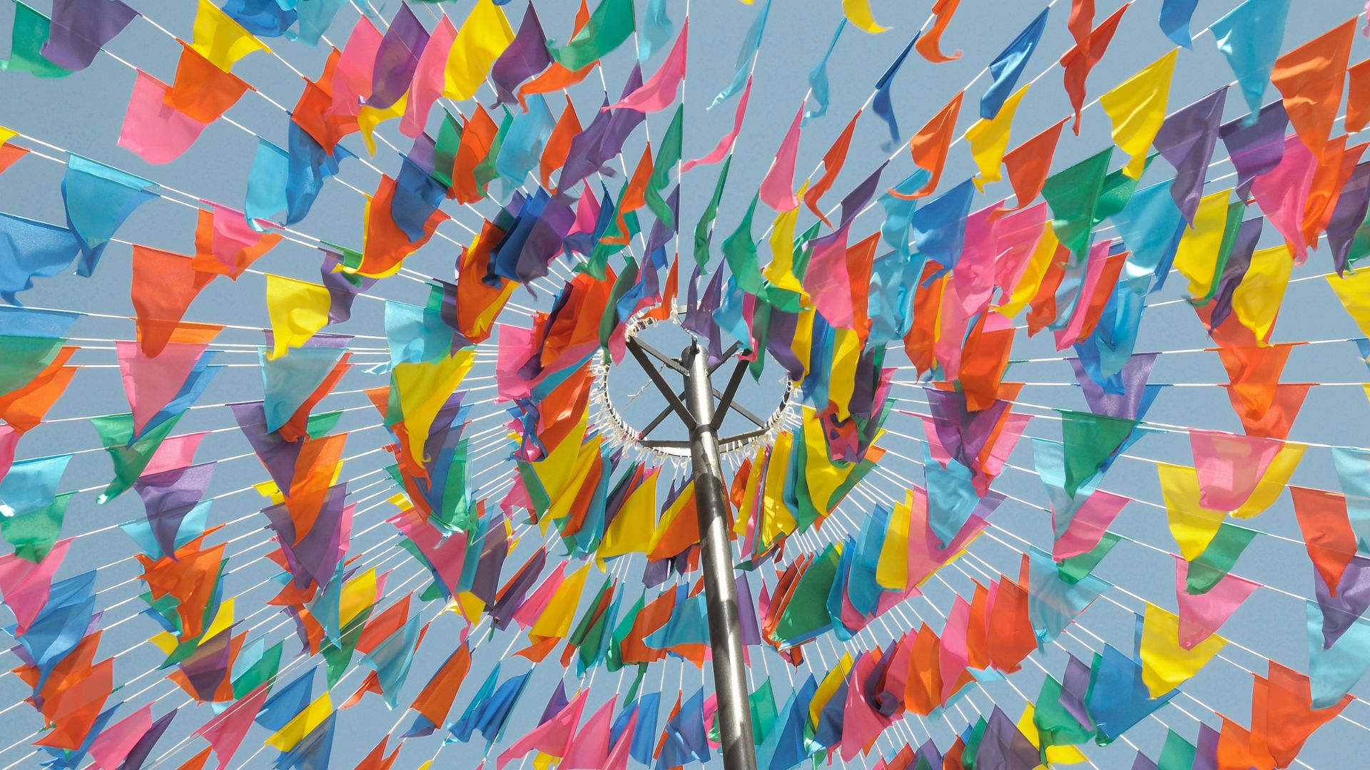 A low-angle view captures numerous colorful triangular flags or pennants strung together, radiating outwards from a central pole against a bright blue sky, creating a festive atmosphere. 