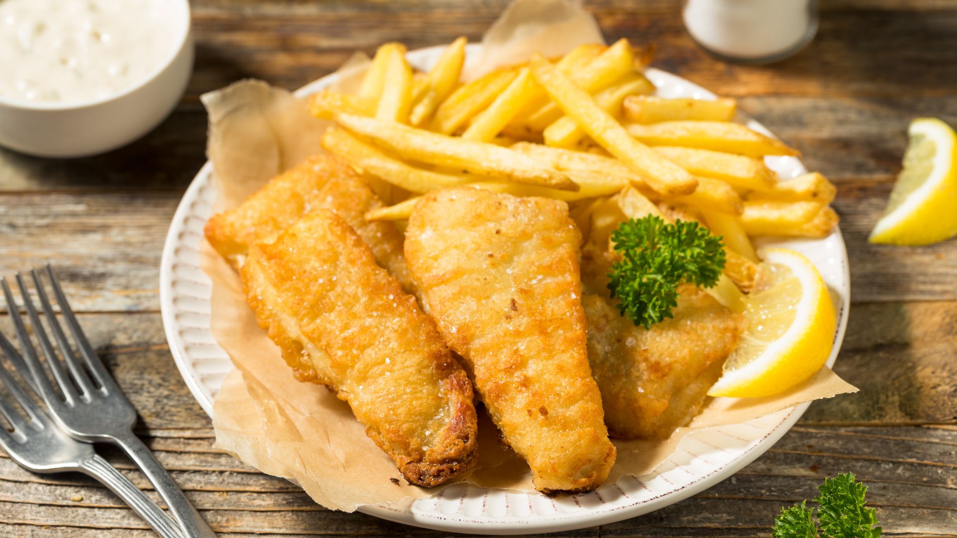 A plate of golden-brown battered fish fillets and a generous serving of crispy french fries, garnished with a lemon wedge and fresh parsley, with a bowl of tartar sauce in the background.