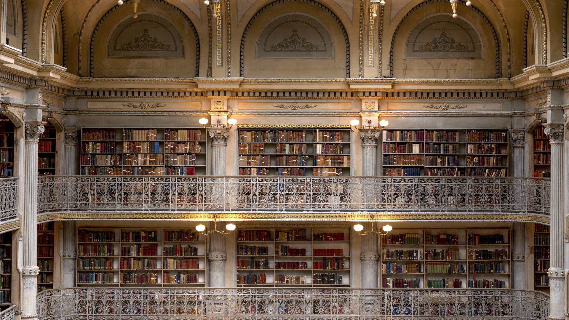 A grand, multi-tiered library interior with ornate cast-iron balconies and towering bookshelves filled with books, reaching up to a skylight, showcasing the architectural beauty of the George Peabody Library.
