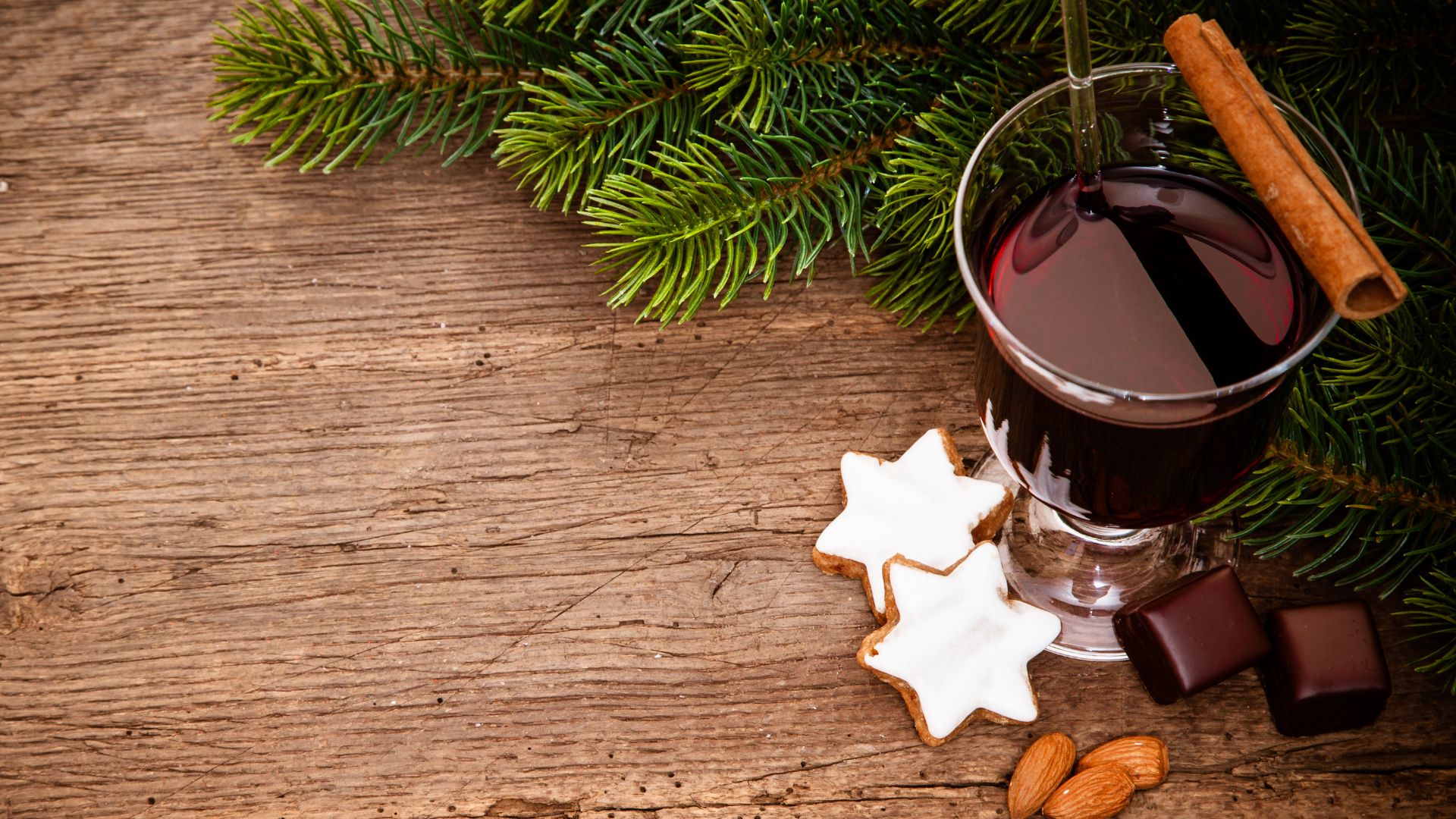 A high-angle shot shows a glass of dark red mulled wine with a cinnamon stick, accompanied by two star-shaped cookies with white icing, two pieces of dark chocolate, and a small pile of almonds, all resting on a wooden surface with green pine needles in the background.