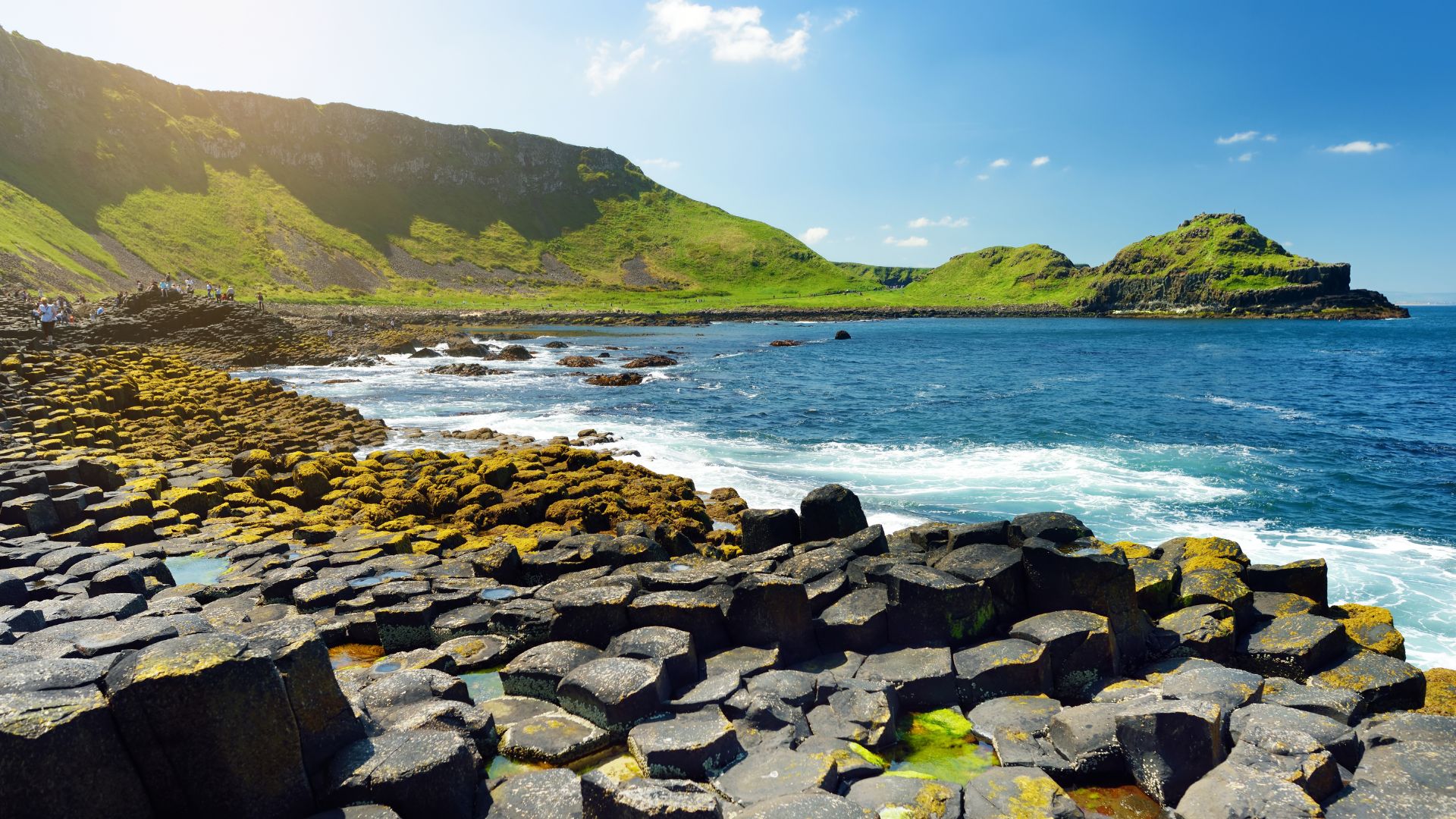 A wide-angle view of the Giant's Causeway, featuring thousands of dark, hexagonal basalt columns forming a rugged coastline leading into the blue sea, with a green, rolling hill under a bright sky in the background.