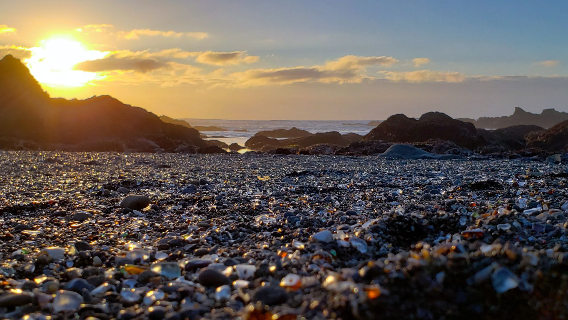 A close-up view of Glass Beach in California, showing a vibrant collection of smoothed, colorful sea glass pebbles mixed with sand and small rocks on the shore, with gentle ocean waves breaking in the background under a clear sky.