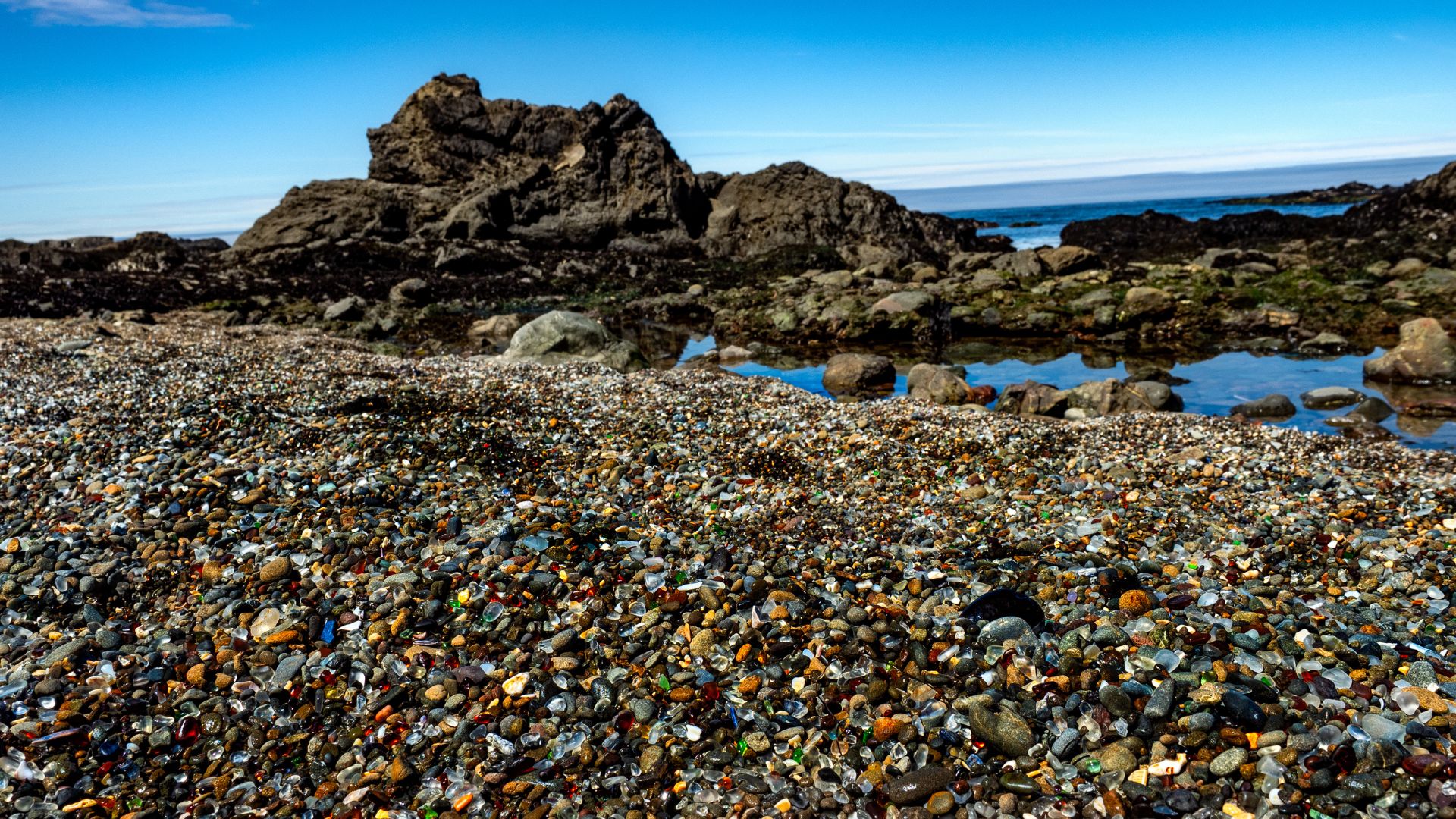 A close-up view of Glass Beach in California, showing a vibrant collection of smoothed, colorful sea glass pebbles mixed with sand and small rocks on the shore, with gentle ocean waves breaking in the background under a clear sky.