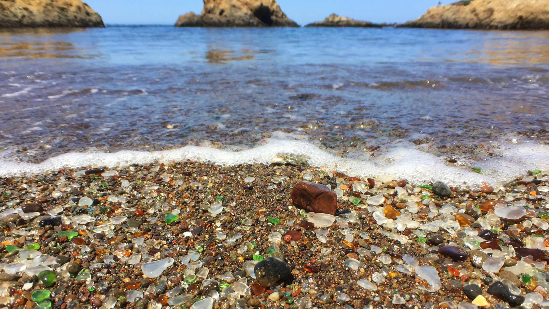A close-up view of Glass Beach in California, showing a vibrant collection of smoothed, colorful sea glass pebbles mixed with sand and small rocks on the shore, with gentle ocean waves breaking in the background under a clear sky.