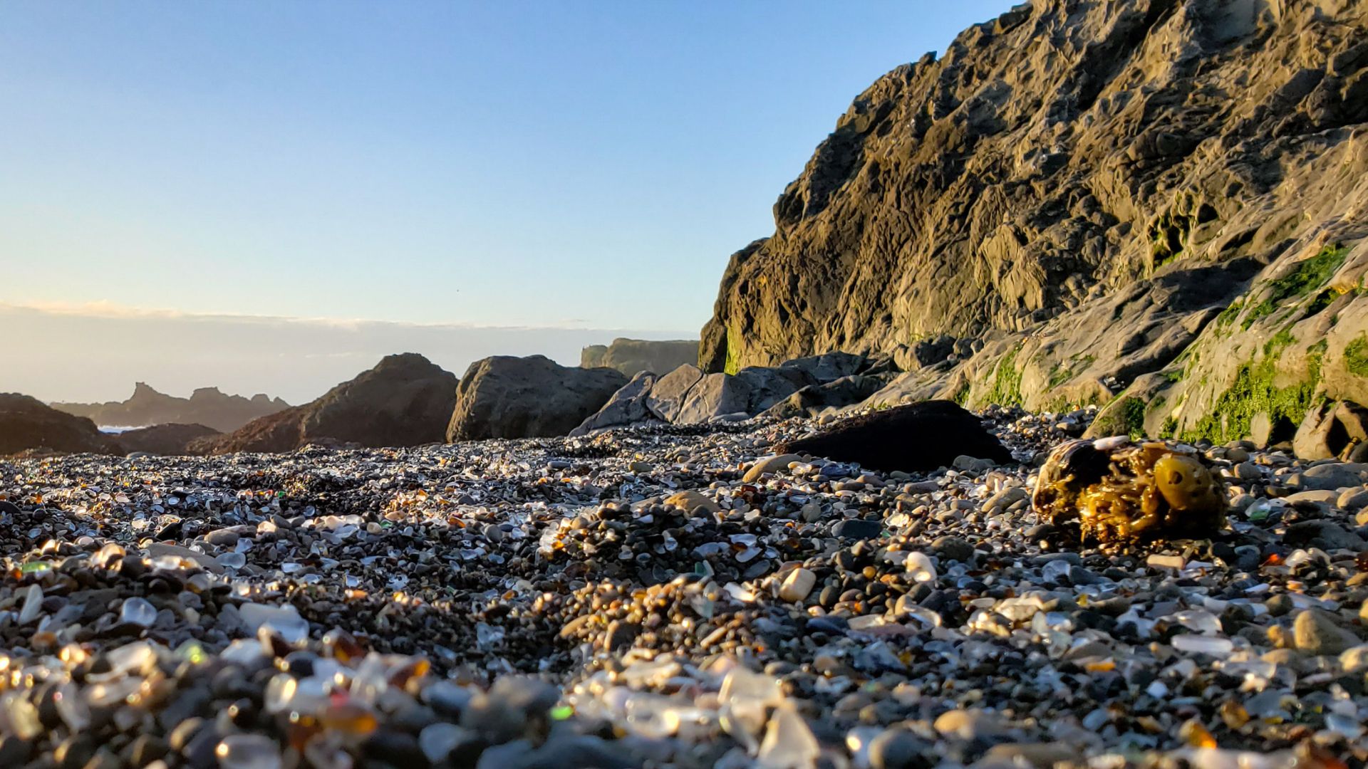 A low-angle shot of Glass Beach in Fort Bragg, California, at sunset, showing a shoreline covered in smooth, colorful sea glass pebbles with a large rocky cliff face on the right and other rock formations in the distance under a clear sky.