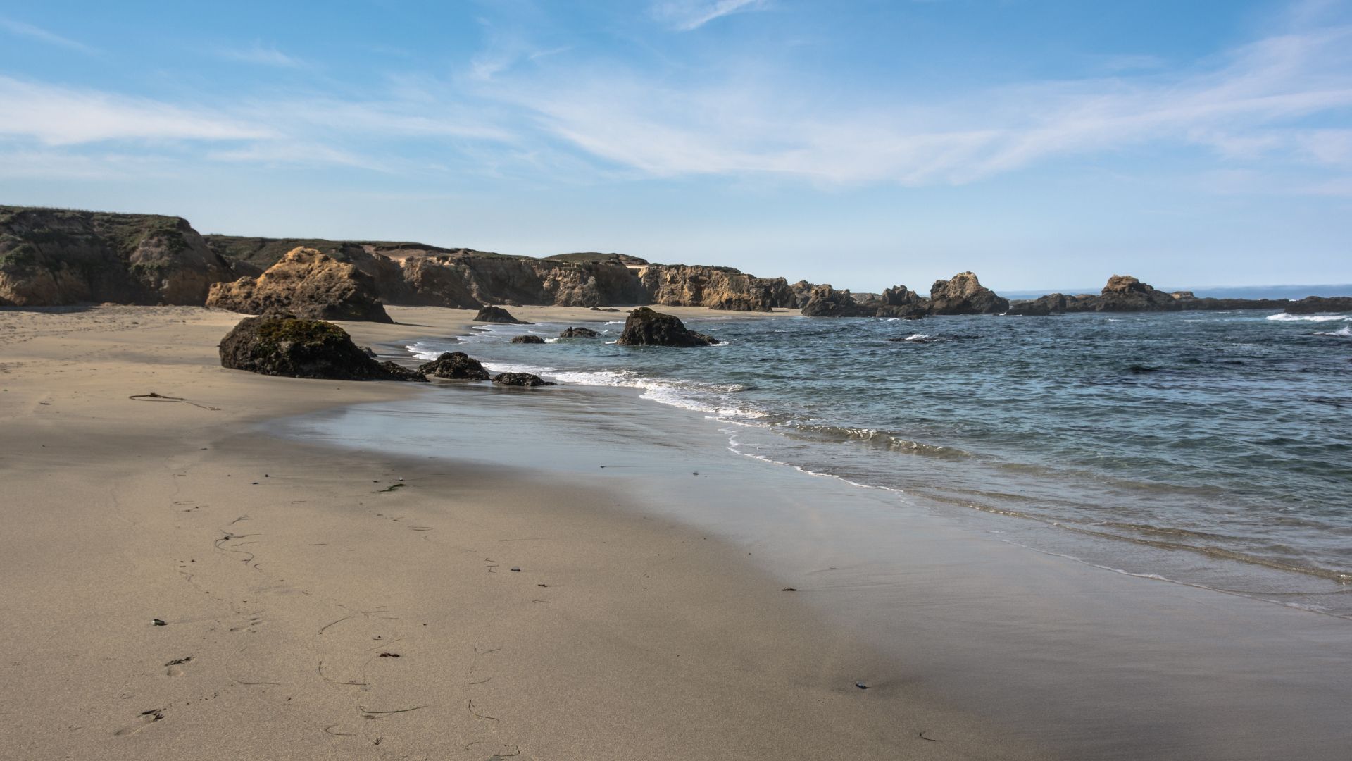 A wide shot of a sandy beach with gentle waves lapping the shore, backed by rocky cliffs under a clear blue sky with scattered clouds.
