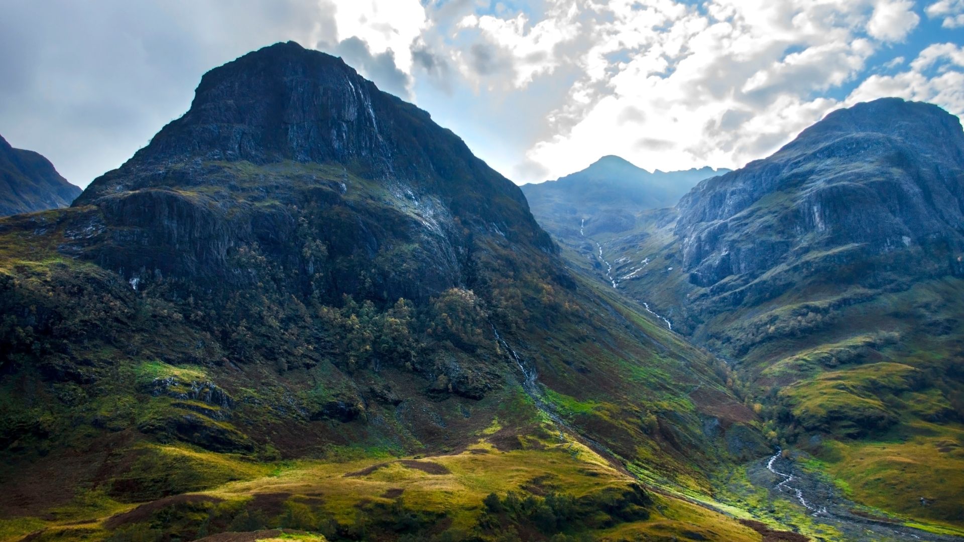 A dramatic landscape photograph of Glen Coe in the Scottish Highlands, featuring rugged, dark mountains covered with patches of green and golden-brown vegetation, a winding stream, and a partially cloudy sky.