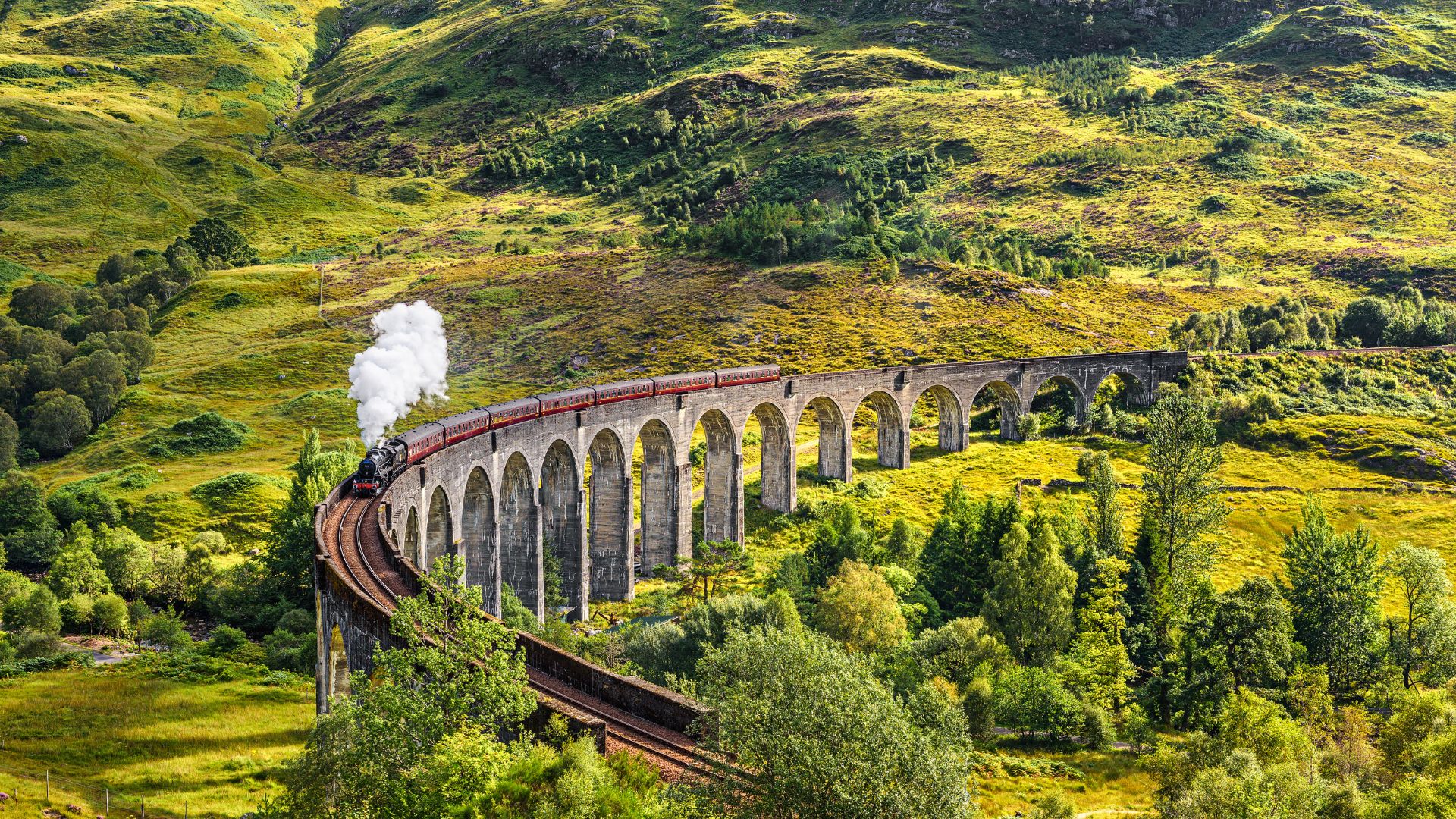A blue and yellow train crosses the arched Glenfinnan Viaduct, a large railway bridge set amidst lush green hills in the Scottish Highlands.