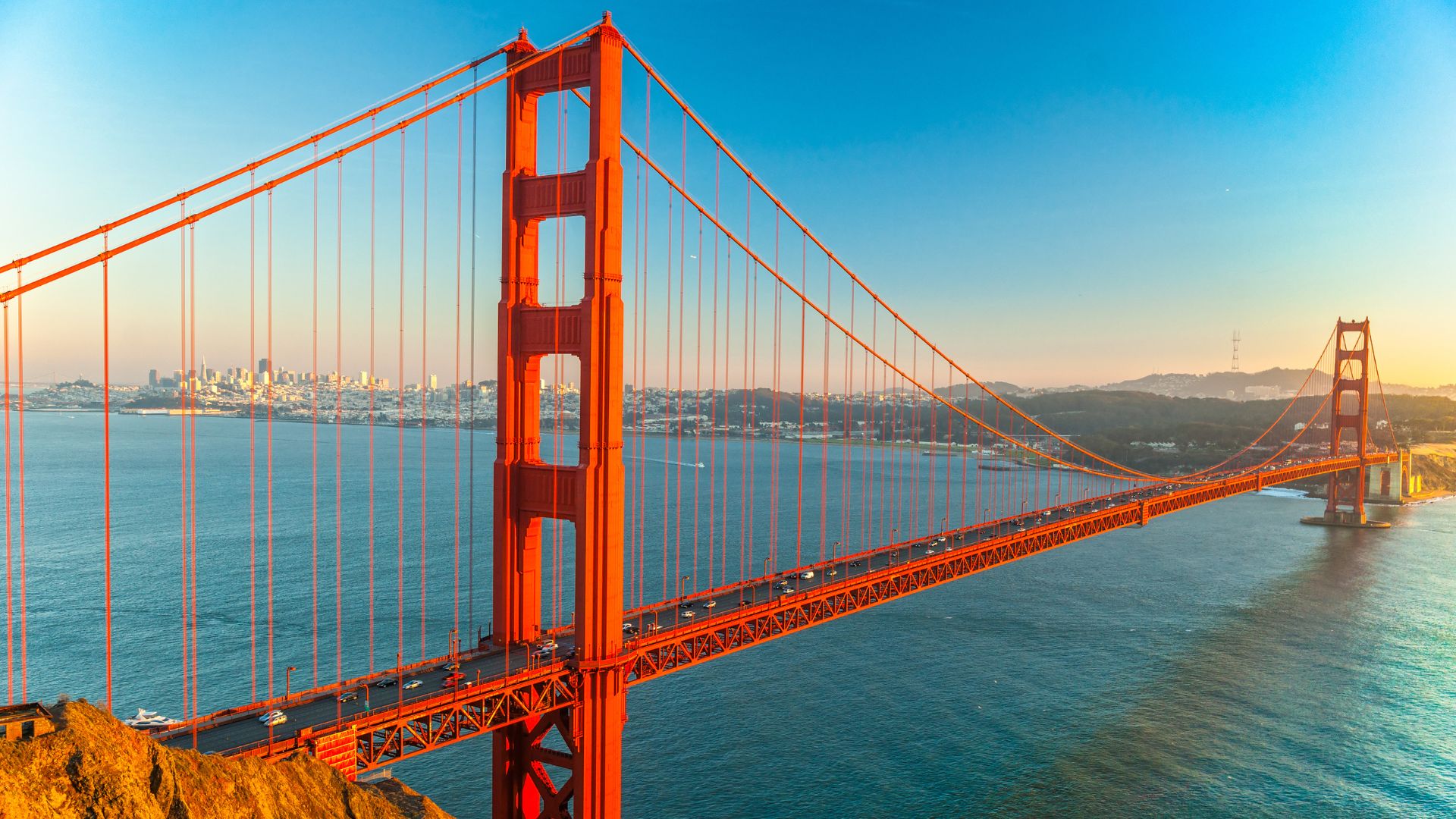 A wide shot of the iconic Golden Gate Bridge, a large red suspension bridge, spanning across a body of water with a city skyline visible in the background under a clear blue sky.