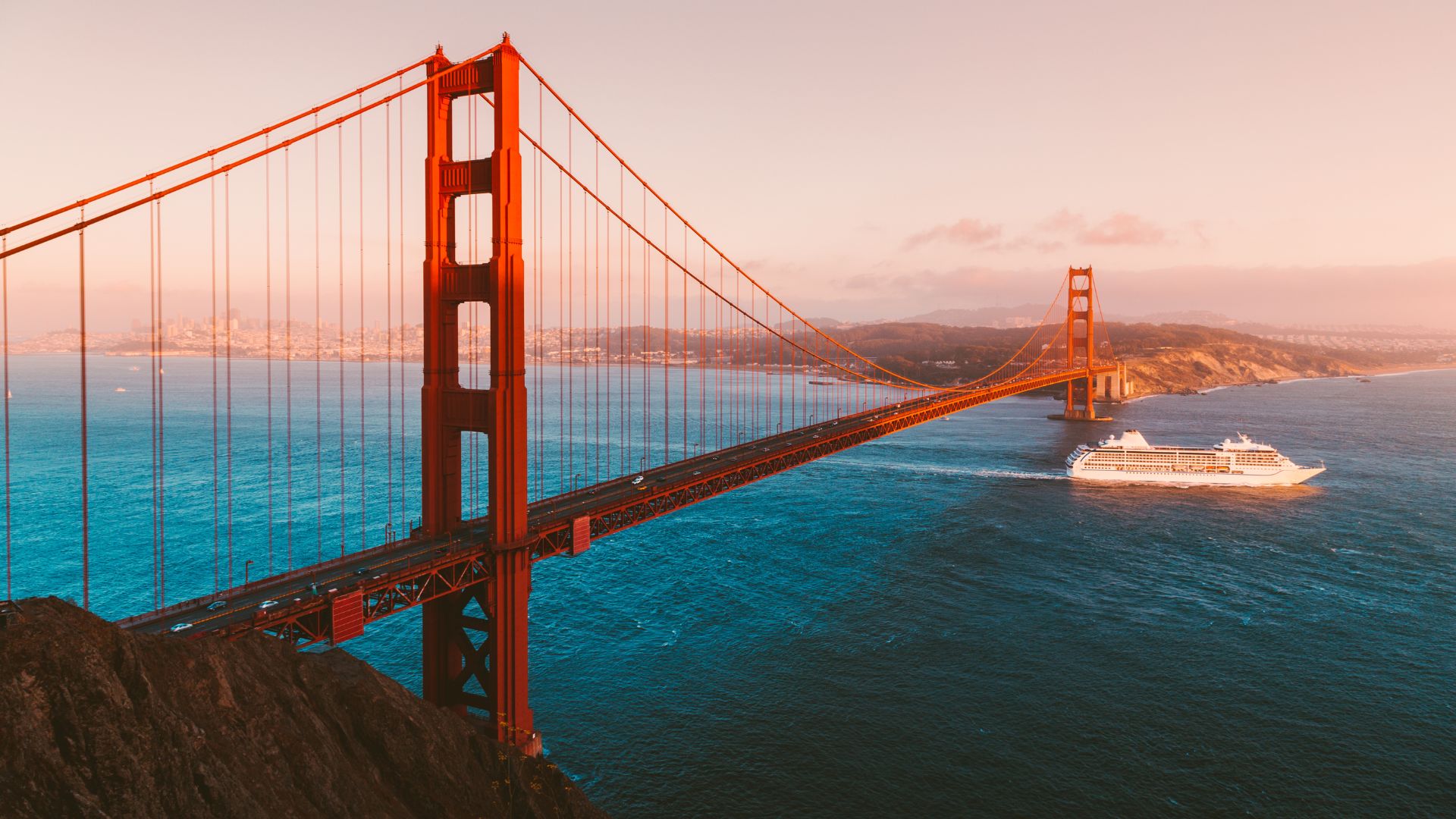 A wide shot of the iconic Golden Gate Bridge, a vibrant orange suspension bridge, spanning across a body of water with a large white cruise ship sailing beneath it towards the right, under a clear sky with a soft, warm light.
