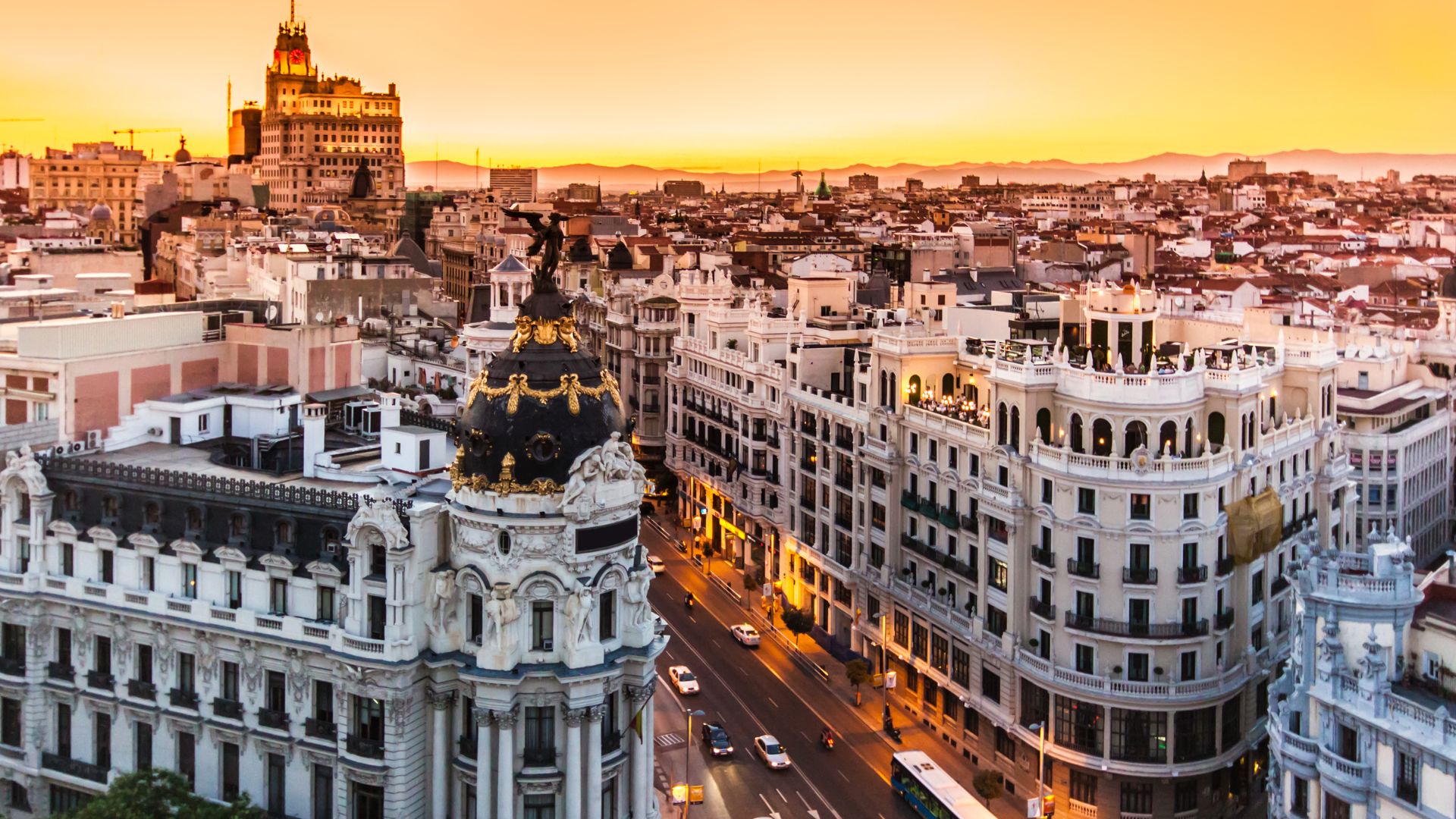 A high-angle view of Madrid's Gran Vía at sunset, showcasing the iconic Metropolis Building and other ornate architecture along the bustling street, with a warm orange glow in the sky. 