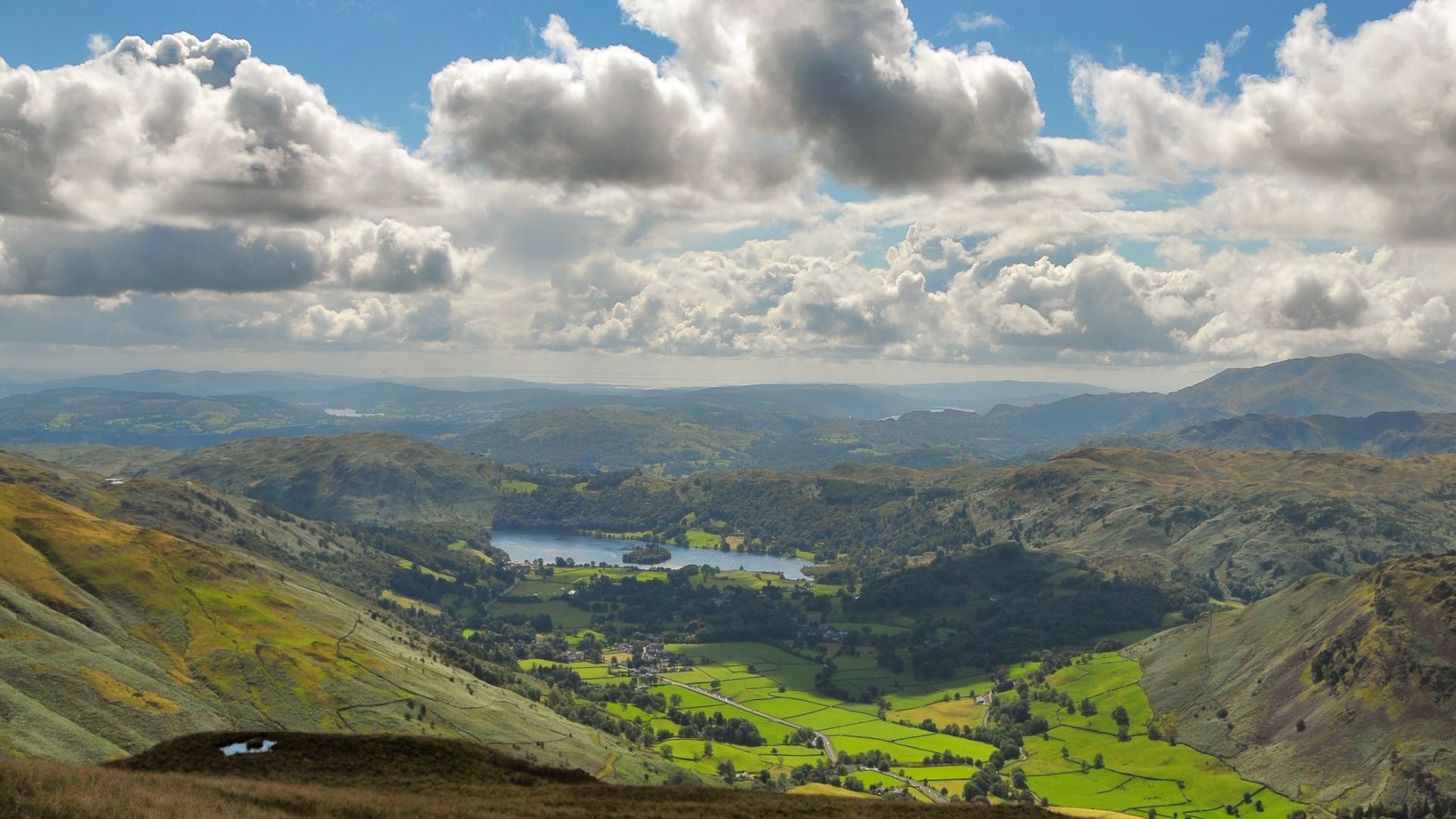 A panoramic view of Grasmere Lake in the Lake District, England, seen from a high vantage point, with rolling green hills and distant mountains under a cloudy sky.