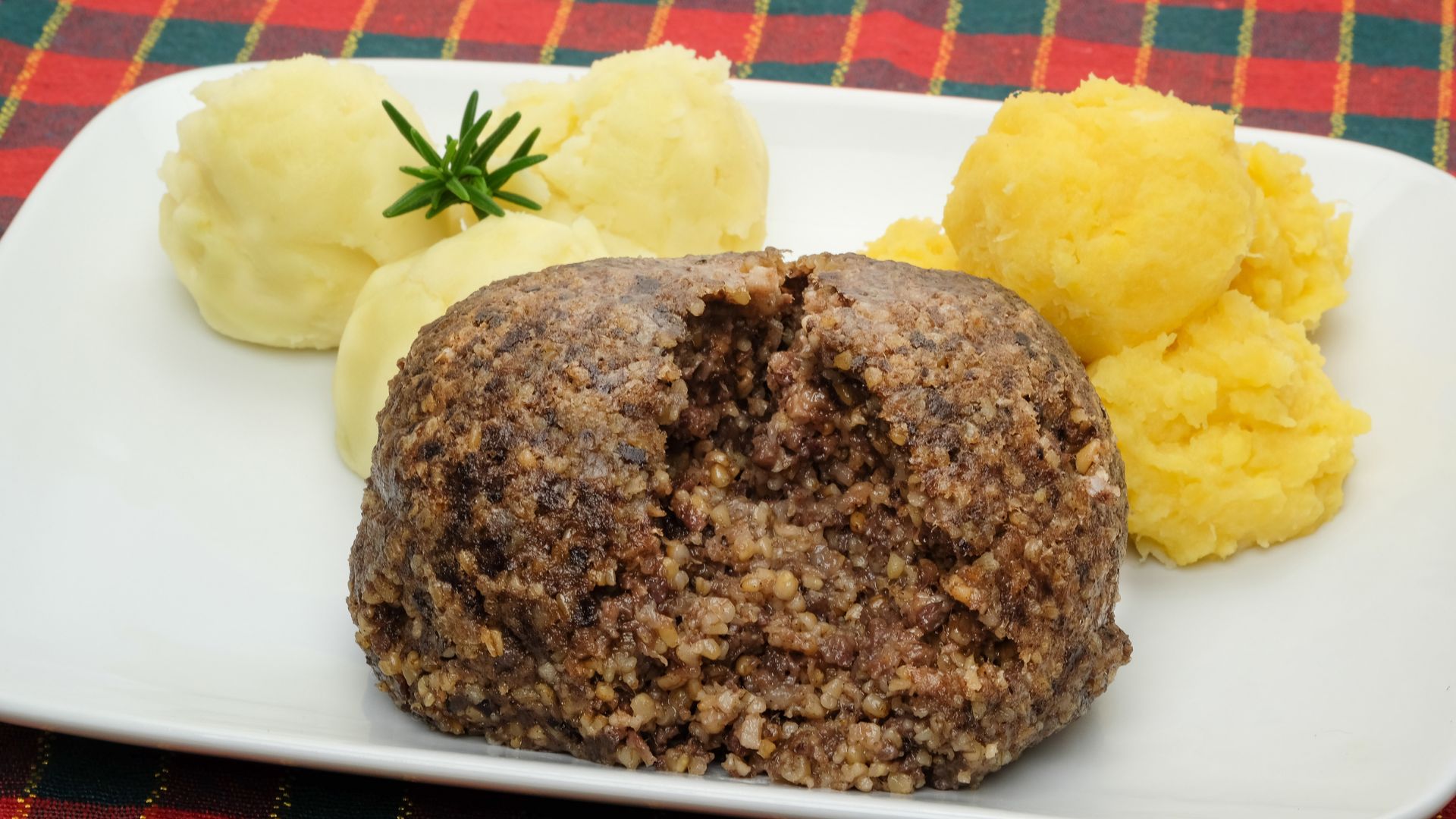 A plate of haggis, a savory pudding, served alongside mashed potatoes ("tatties") and mashed turnips or swede ("neeps"), with a sprig of rosemary for garnish.