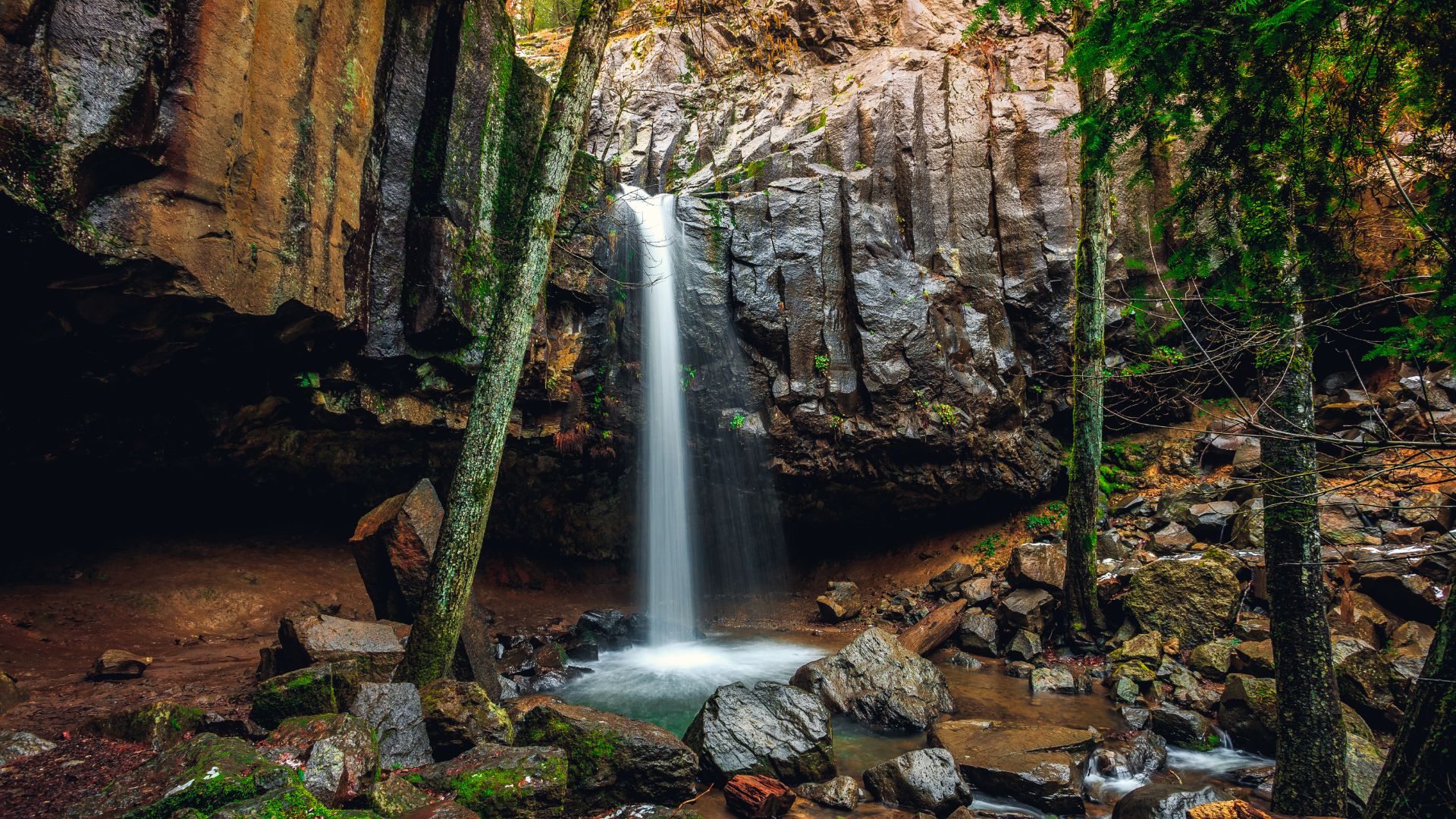 A long exposure photograph captures Hedge Creek Falls cascading down a columnar basalt cliff face into a rocky pool, surrounded by lush green trees and moss-covered rocks, with a visible cave behind the falls. 