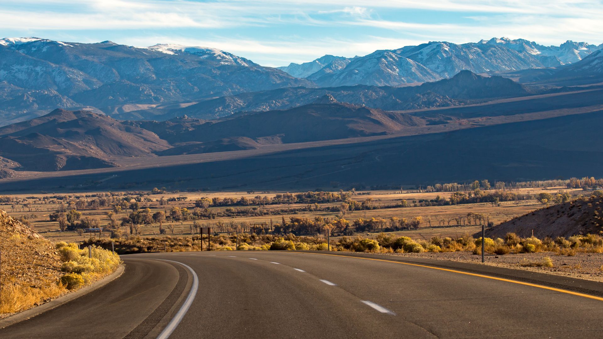 A scenic view of a highway curving through a vast valley with golden fields and sparse trees, leading towards majestic snow-capped mountains under a clear sky.