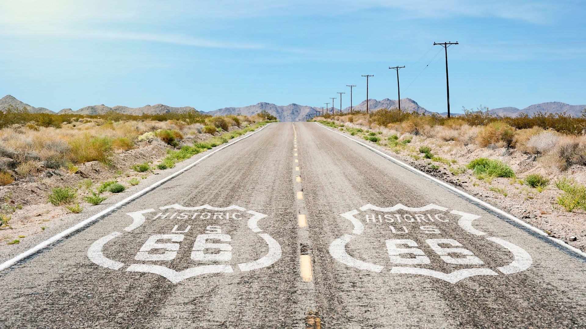 An empty stretch of Historic Route 66 in the Mojave Desert, California, with "Historic US 66" shields painted on the asphalt, leading towards distant mountains under a clear blue sky. Utility poles line the right side of the road.