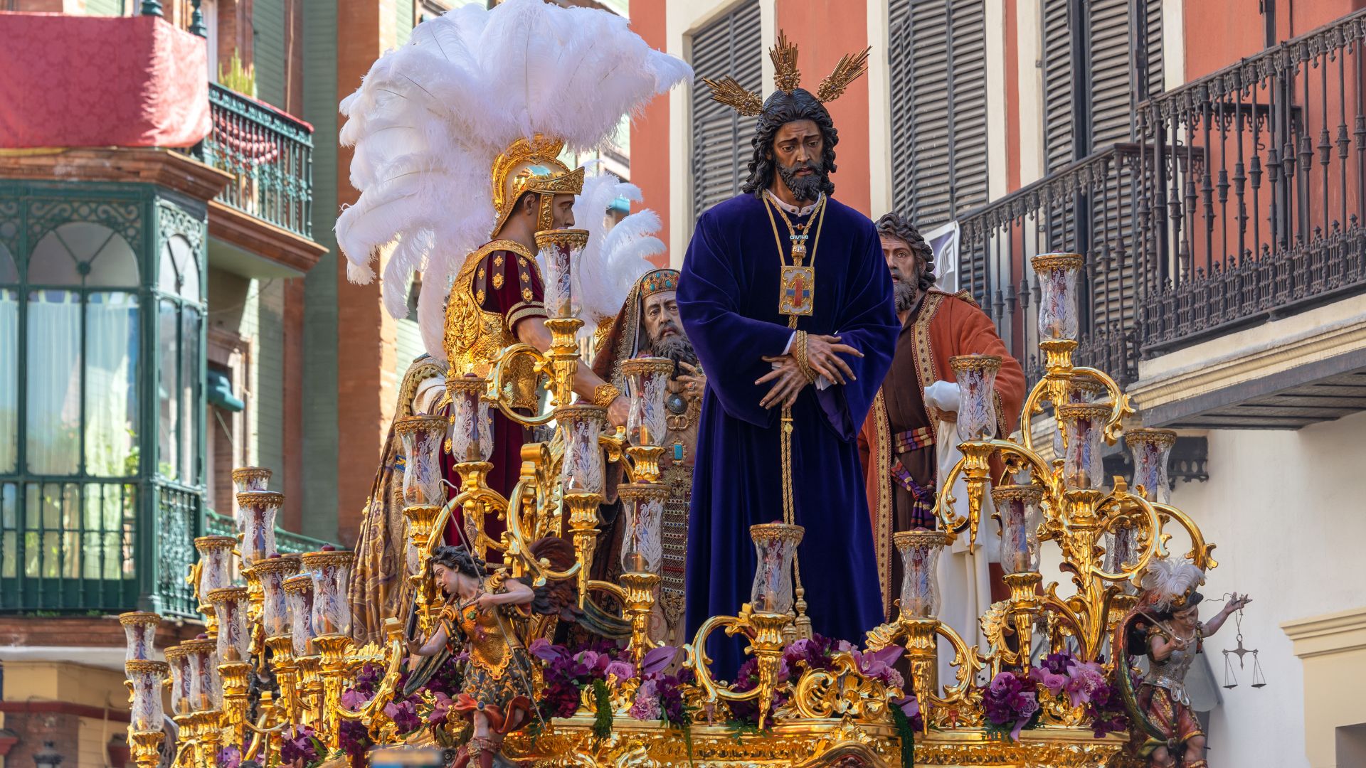 A vibrant street scene during Holy Week in Seville, Spain, showing a large, ornate golden float carrying religious statues, including a prominent figure of Jesus Christ in a blue robe, accompanied by other figures and adorned with candles and flowers, surrounded by buildings with balconies.