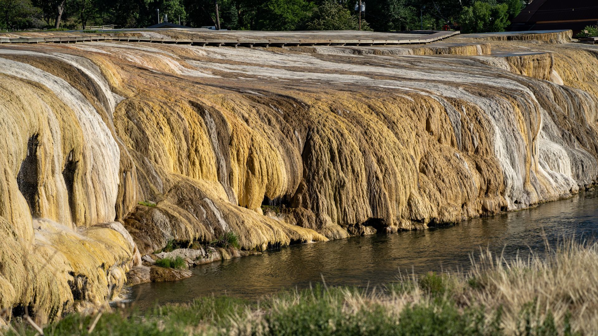 A wide shot shows the vibrant, multi-colored mineral terraces of Hot Springs State Park in Thermopolis, Wyoming, with mineral-rich water flowing over them into a calm river below, flanked by green grasses and a boardwalk visible in the upper background.