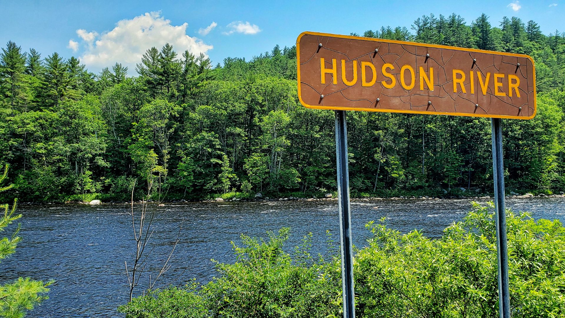 A brown sign with yellow text reading "HUDSON RIVER" stands in the foreground, with the dark waters of the Hudson River and a dense green forest on the opposite bank visible in the background under a blue sky with some clouds.