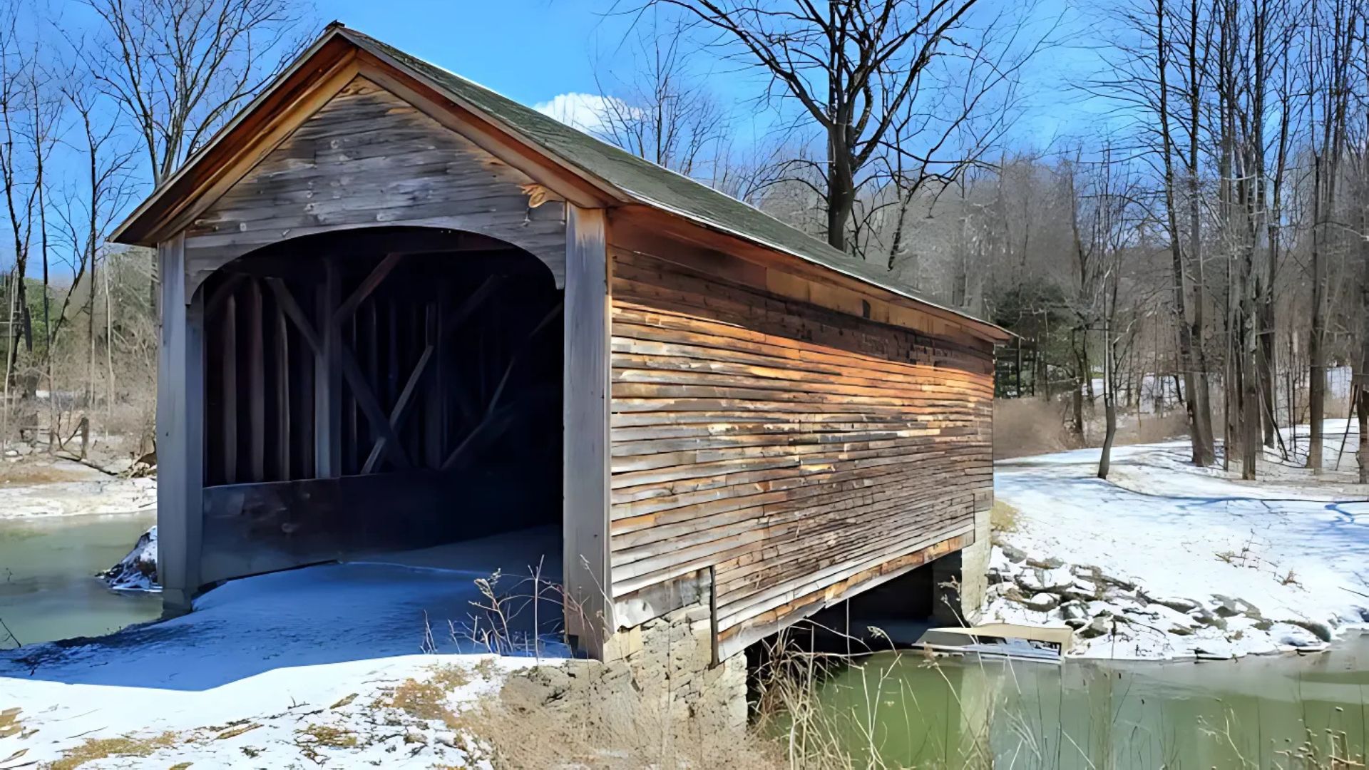 A wooden covered bridge spans a partially snow-covered stream or river, with bare trees and a snowy landscape visible in the background under a clear blue sky.