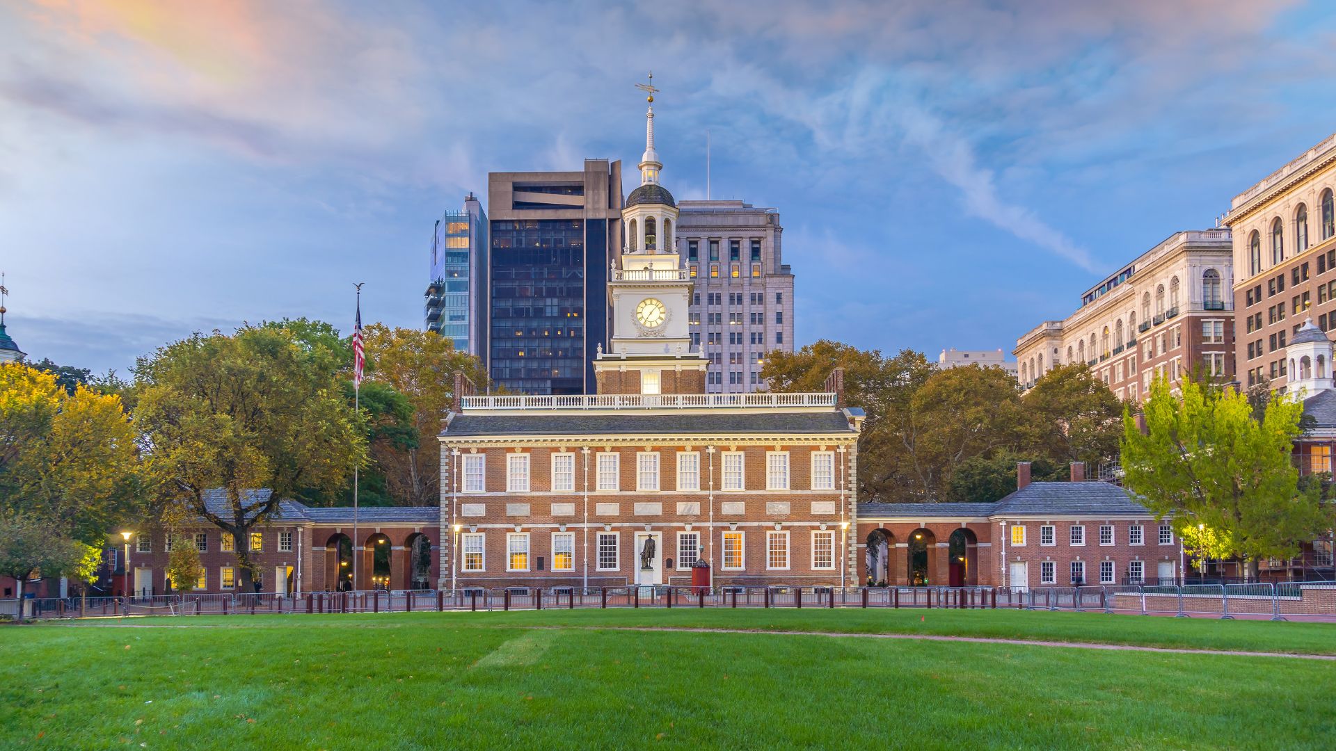 A wide shot of Independence Hall in Philadelphia, Pennsylvania, a historic brick building with a prominent clock tower and steeple, surrounded by green grass and trees under a partly cloudy sky at dusk.