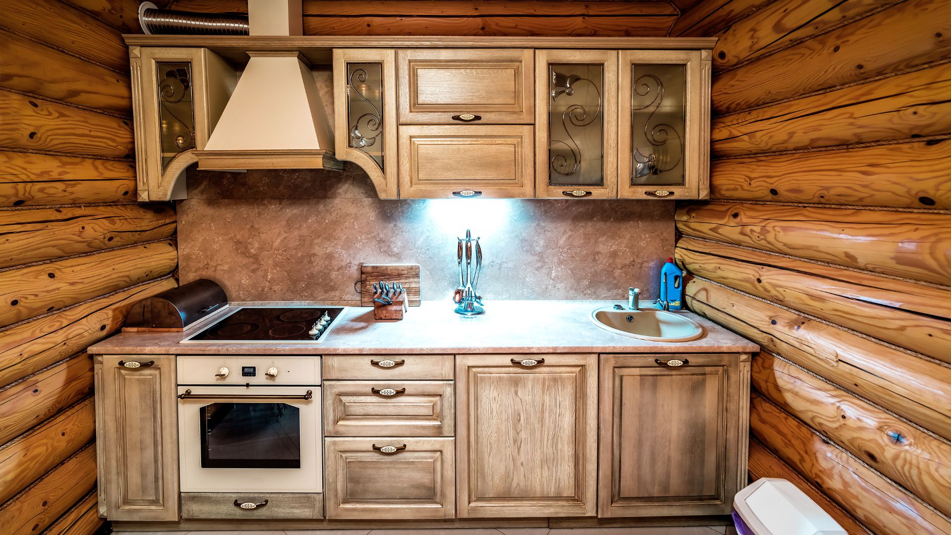 A rustic kitchen in a log cabin features light-colored wooden cabinets, a built-in oven and stovetop, a white countertop with a sink, and exposed log walls. 