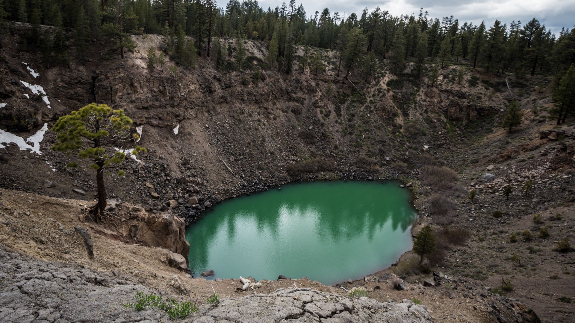 A deep, funnel-shaped volcanic crater with an emerald-green lake at its base, surrounded by rocky slopes and sparse trees under a cloudy sky in California.