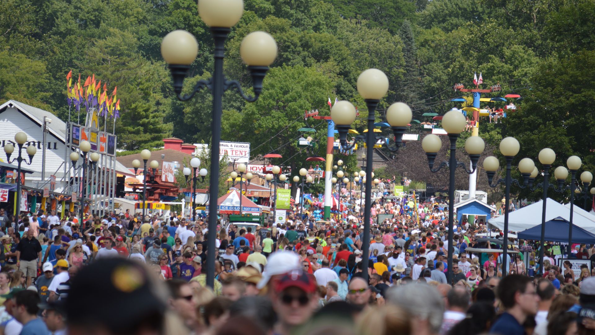 A wide shot of a bustling outdoor fairground filled with a large crowd of people, numerous food stalls, carnival rides, and tall street lamps with round, glowing lights, all set against a backdrop of lush green trees and a clear sky.