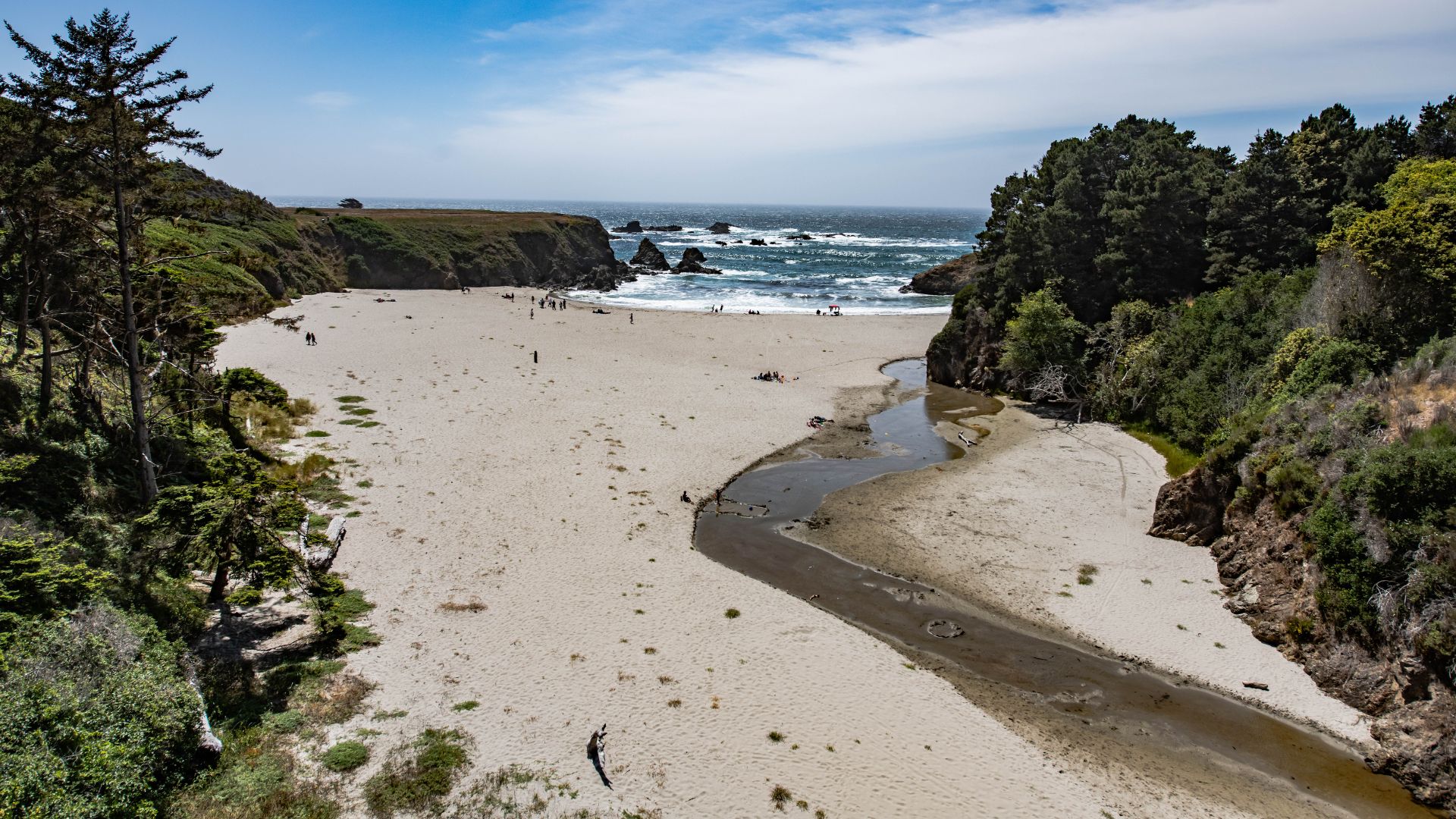 A wide-angle aerial view of a sandy beach cove with a winding creek flowing into the ocean, flanked by lush green bluffs and trees under a clear blue sky.