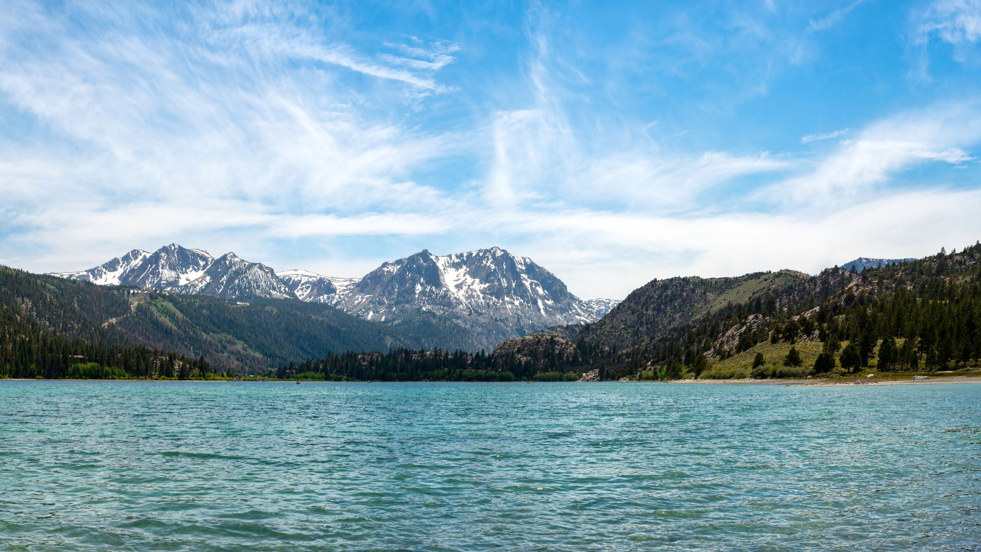 A wide shot of June Lake, a clear blue-green lake, reflecting a bright blue sky with wispy clouds. Snow-capped mountains rise majestically in the background, with pine forests lining the lake's shores and the lower mountain slopes.