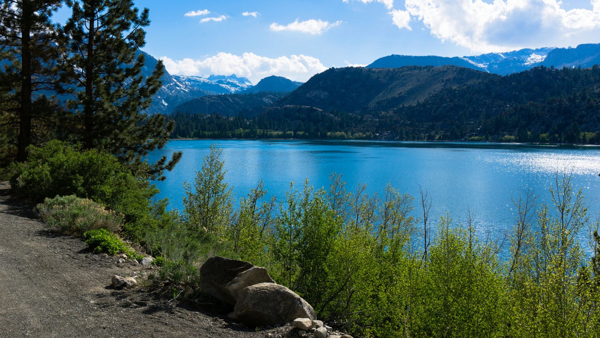A serene landscape photograph featuring a bright blue lake surrounded by lush green trees and shrubs in the foreground, with snow-capped mountains under a partly cloudy sky in the background. A dirt path runs along the left side of the lake.
