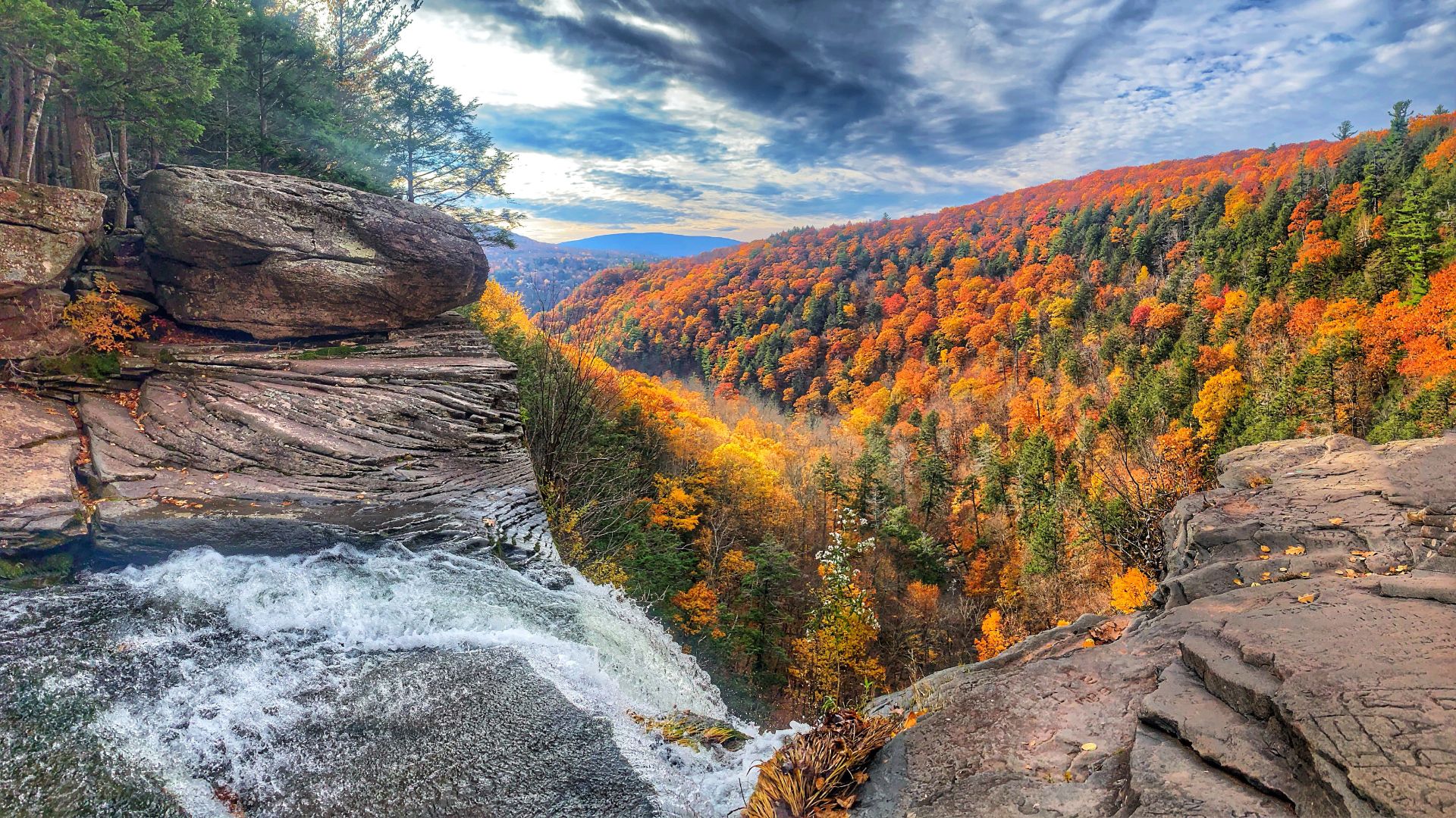 A vibrant autumn landscape featuring Kaaterskill Falls, a two-tiered waterfall cascading down a rocky cliff face into a valley filled with trees displaying brilliant orange and green fall foliage under a dramatic sky.