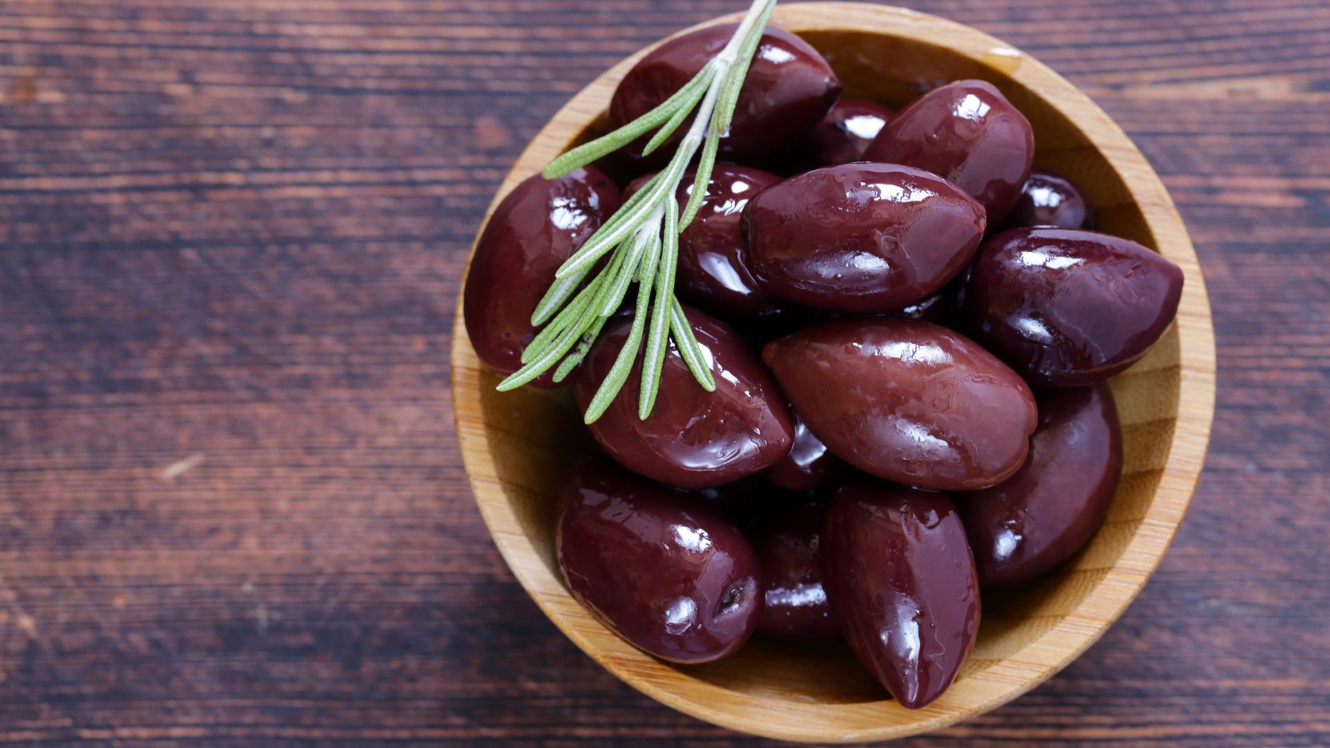 A wooden bowl filled with dark, glossy Kalamata olives and a fresh sprig of rosemary on a rustic wooden surface.