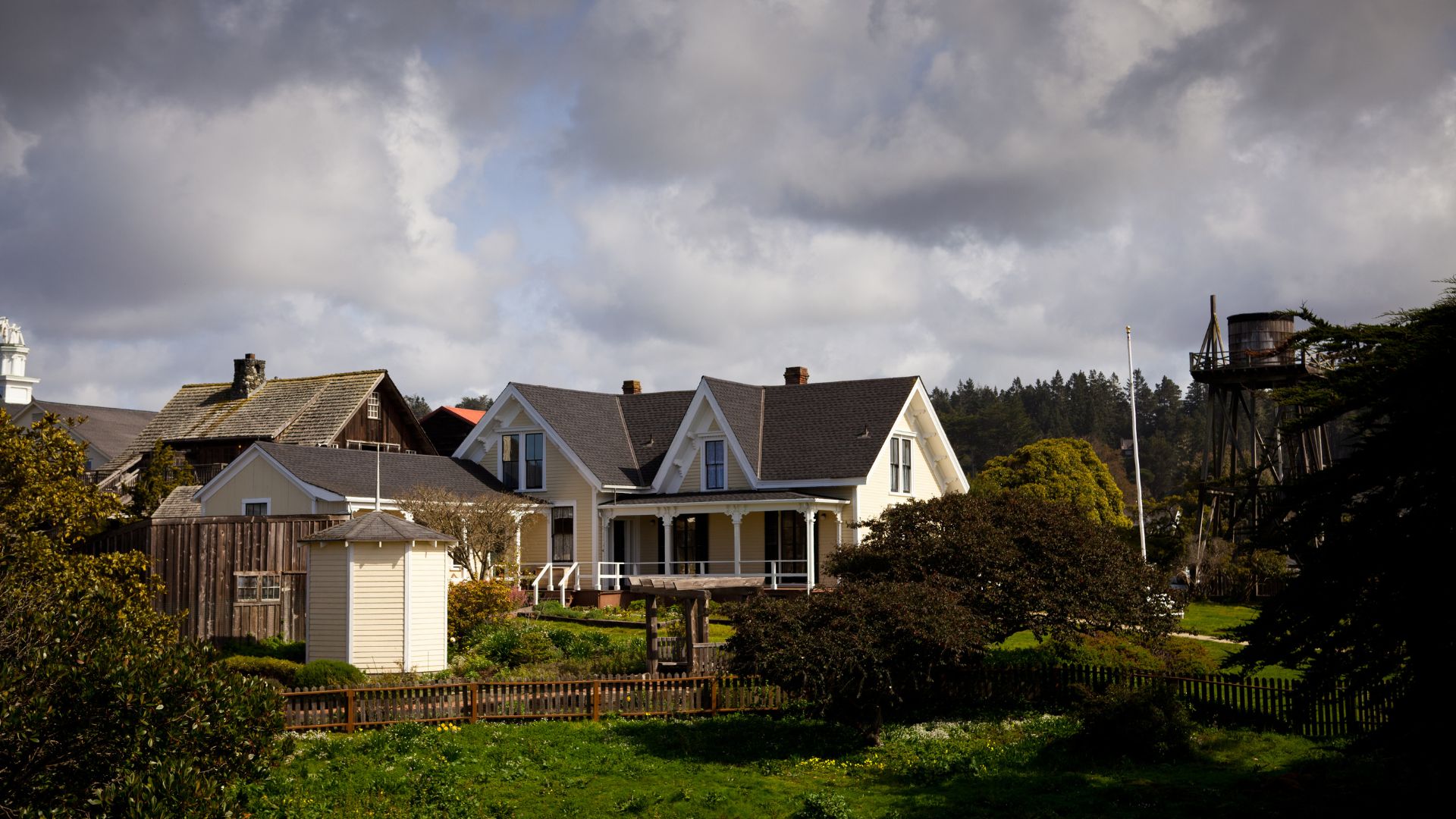 A panoramic view of the Kelley House Museum in Mendocino, California, featuring a historic two-story house with a porch and surrounding gardens, with other buildings and a water tower visible in the background against a cloudy sky.