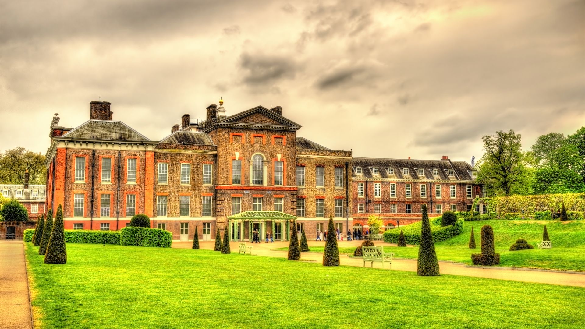 A grand, historic palace with a red-brick facade and classical architectural elements, surrounded by expansive green lawns with manicured hedges and conical topiaries under a cloudy sky.