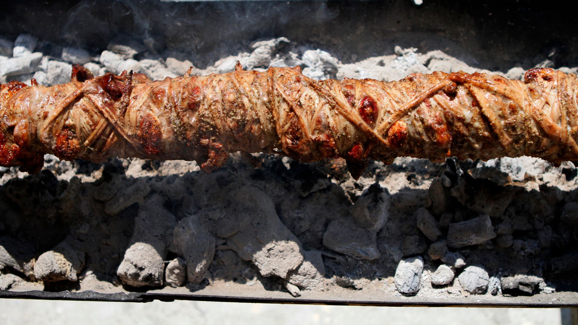 A large, cylindrical roll of seasoned lamb or goat offal wrapped in intestines, known as Kokoretsi, roasting on a spit over hot coals.