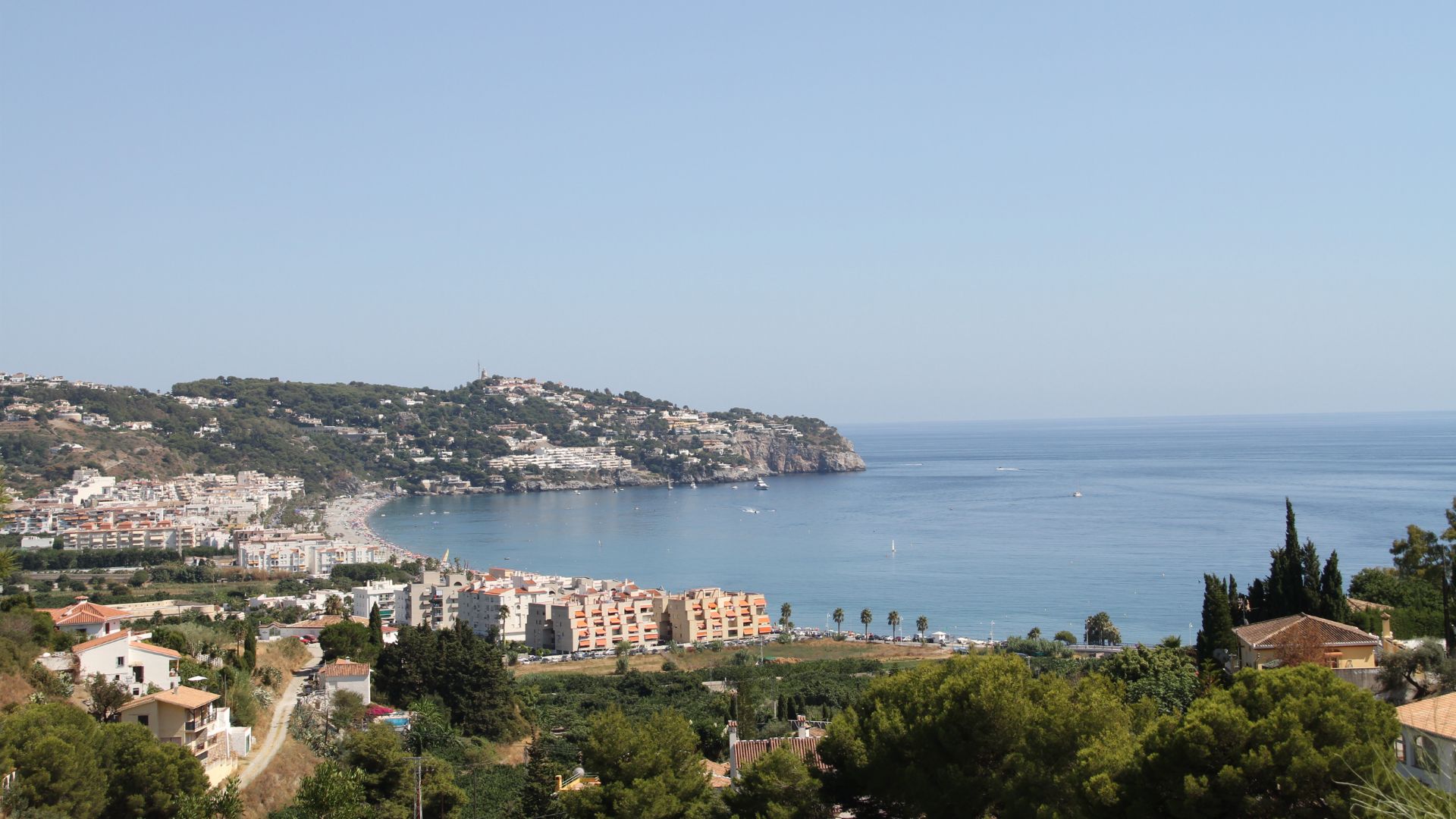 A panoramic view of La Herradura bay in Spain, showing a horseshoe-shaped bay with a beach, coastal town, and buildings nestled among green hills, with the Mediterranean Sea stretching out under a clear sky.