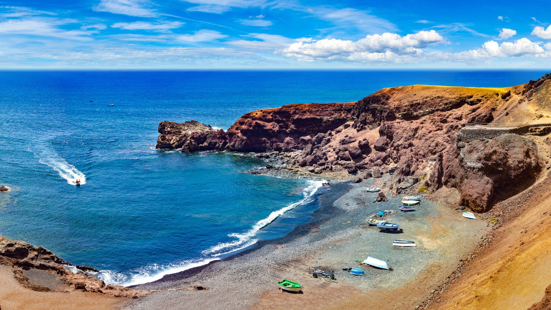 A high-angle view of a volcanic black sand beach with small boats pulled ashore, framed by dramatic dark cliffs on the right and clear blue ocean with a small boat leaving a wake on the left, under a bright blue sky with scattered clouds.