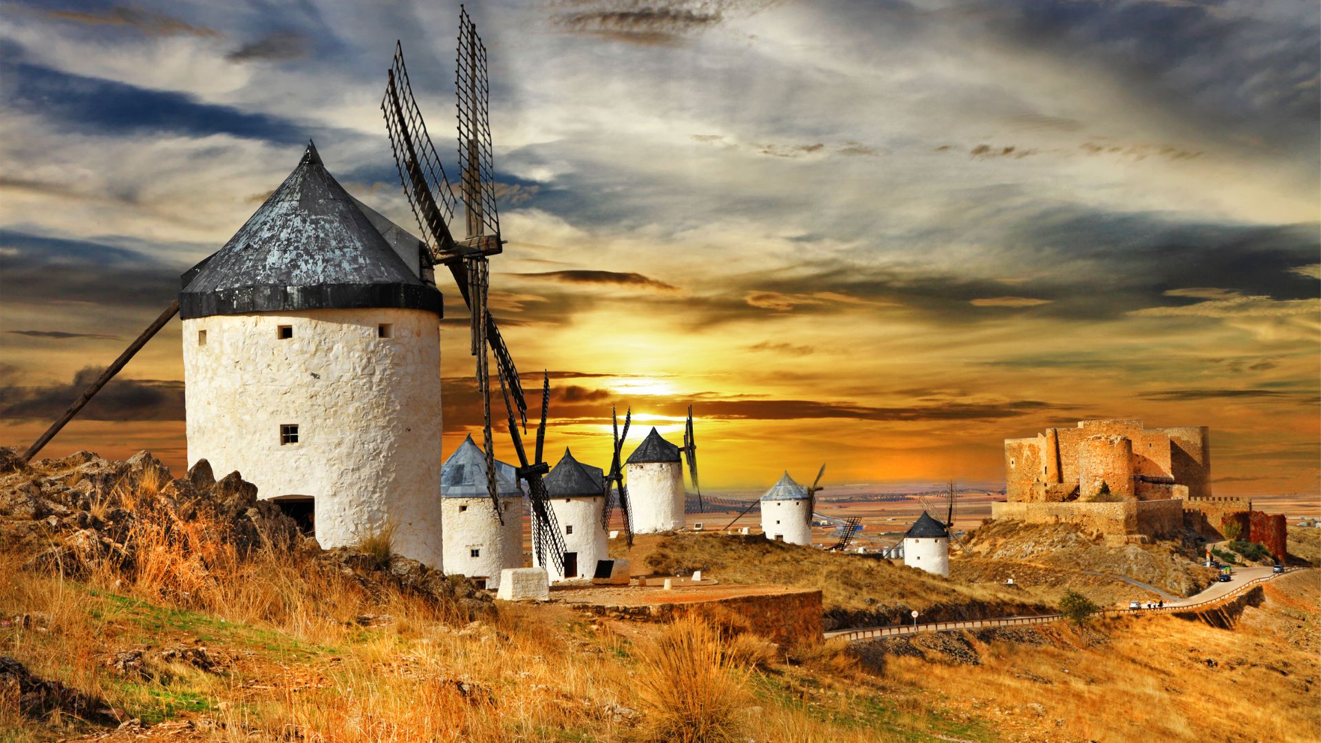 A panoramic view at sunset of the historic Consuegra windmills and a castle on a hill in Castilla-La Mancha, Spain. Several white stone windmills with dark conical roofs and prominent blades are scattered across a dry, grassy landscape, with a large, ancient stone castle visible on a higher point to the right. The sky is filled with dramatic clouds illuminated by the warm glow of the setting sun.
