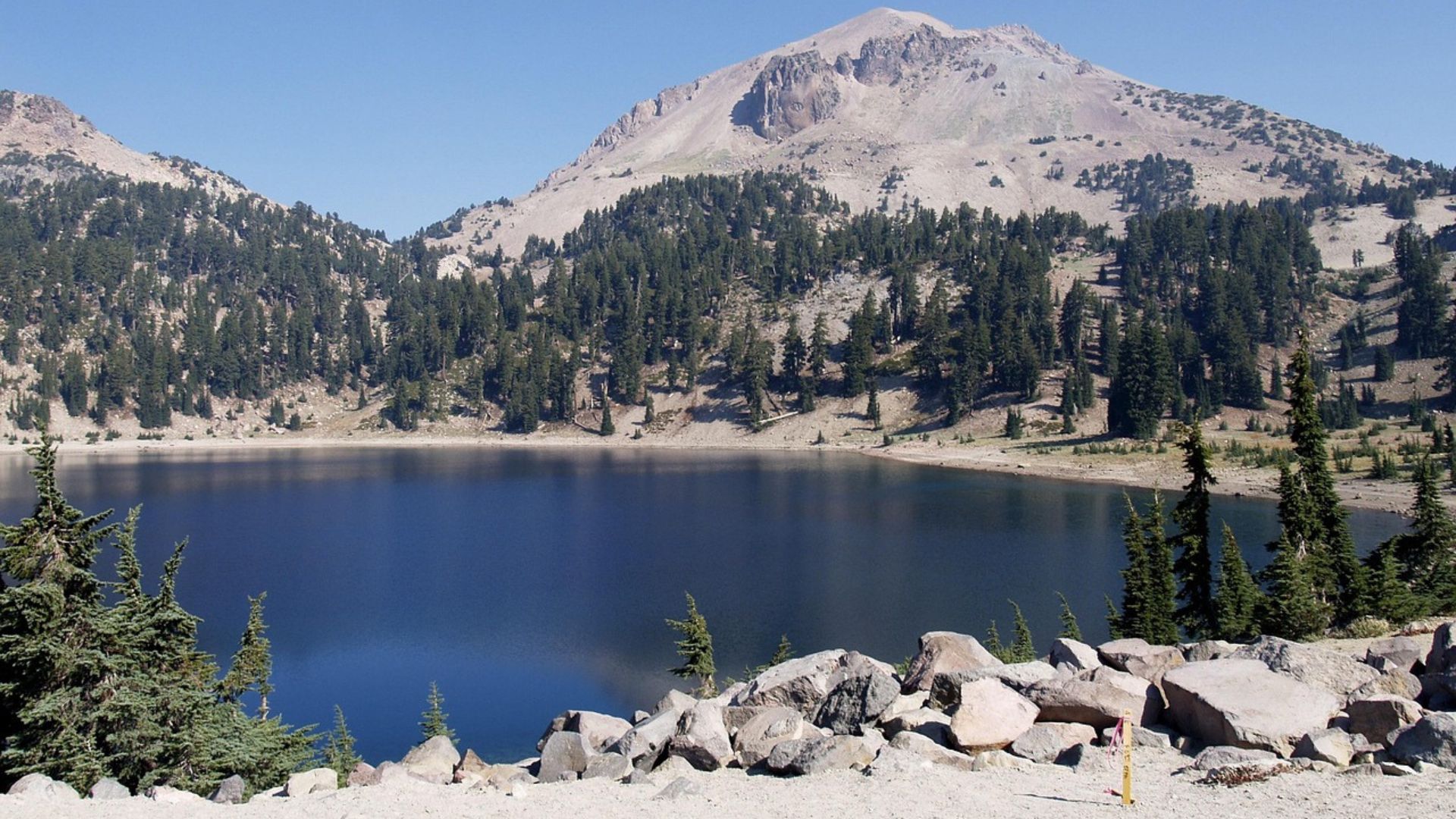 A clear blue lake, Lake Helen, sits in the foreground with a rocky shore, reflecting the surrounding evergreen-covered mountains and the prominent Lassen Peak under a bright blue sky.