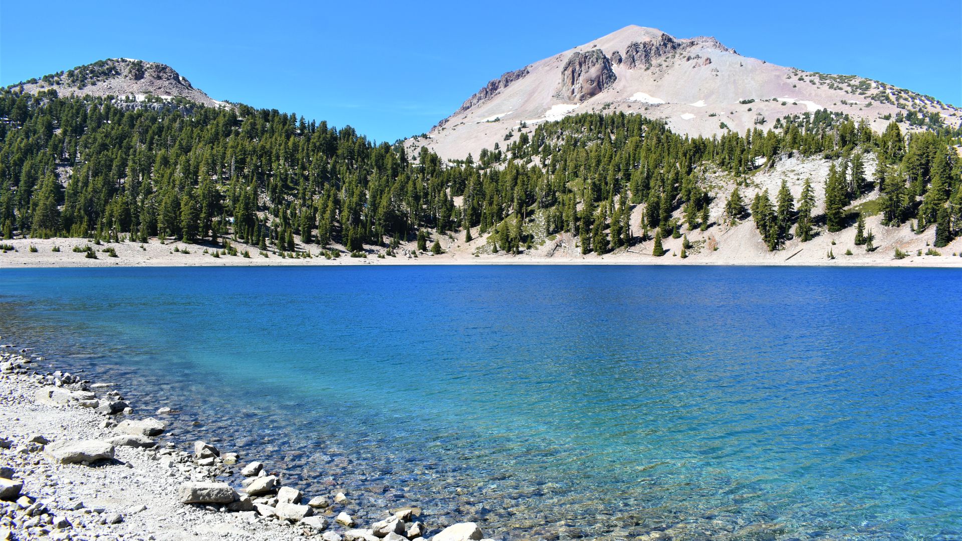 A clear blue lake, Lake Helen, in the foreground with a rocky shoreline, leading to a dense forest of evergreen trees and majestic, snow-dusted mountains, including Lassen Peak, under a bright blue sky in Lassen Volcanic National Park, California.