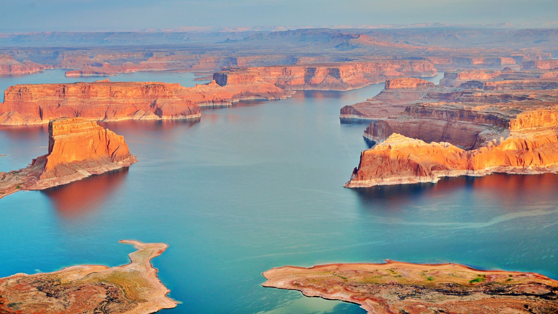An aerial view of Lake Powell, a vast blue lake winding through a landscape of towering orange and red sandstone buttes and canyons under a clear sky.
