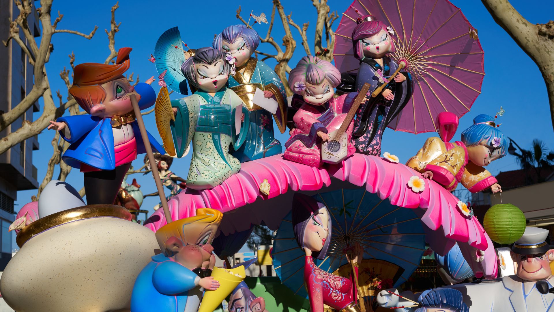 A colorful and ornate "Falla" monument from the Las Fallas Festival in Valencia, Spain, featuring large, sculpted figures in traditional attire, one holding an umbrella, set against a bright blue sky.