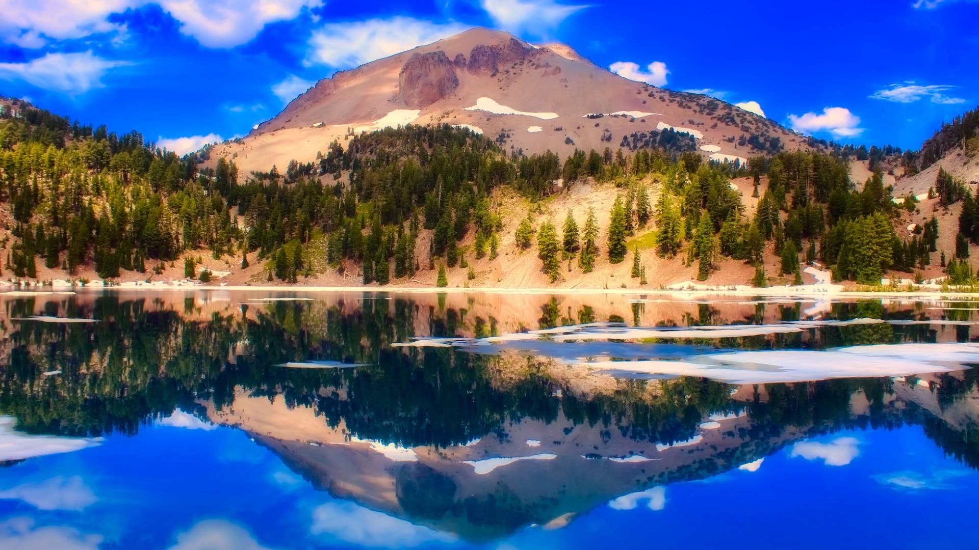 A vibrant landscape photo shows Lassen Peak, a large mountain with patches of snow and surrounded by evergreen trees, perfectly reflected in the calm, blue waters of a lake under a bright blue sky with scattered clouds.