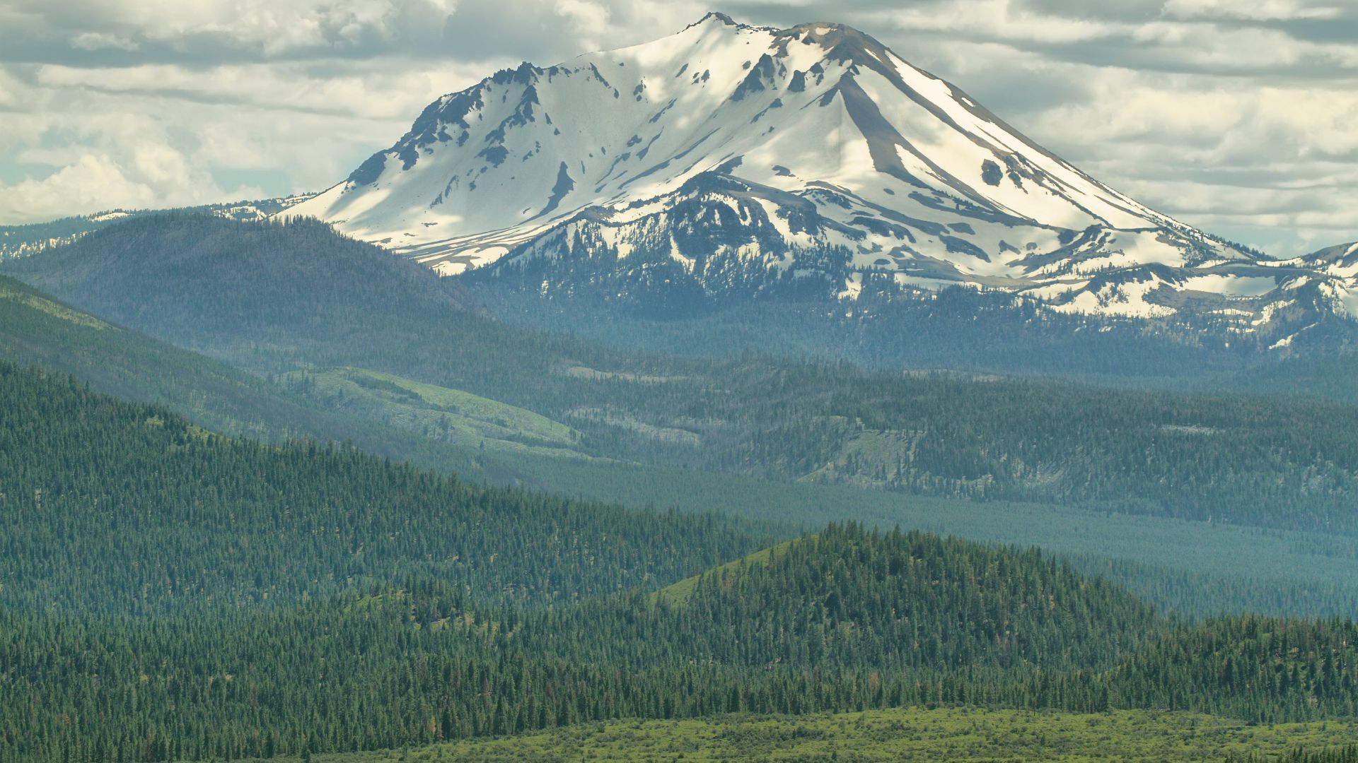 A snow-capped Lassen Peak, a large volcanic mountain, rises above a vast expanse of lush green forest under a partly cloudy sky in Northern California.