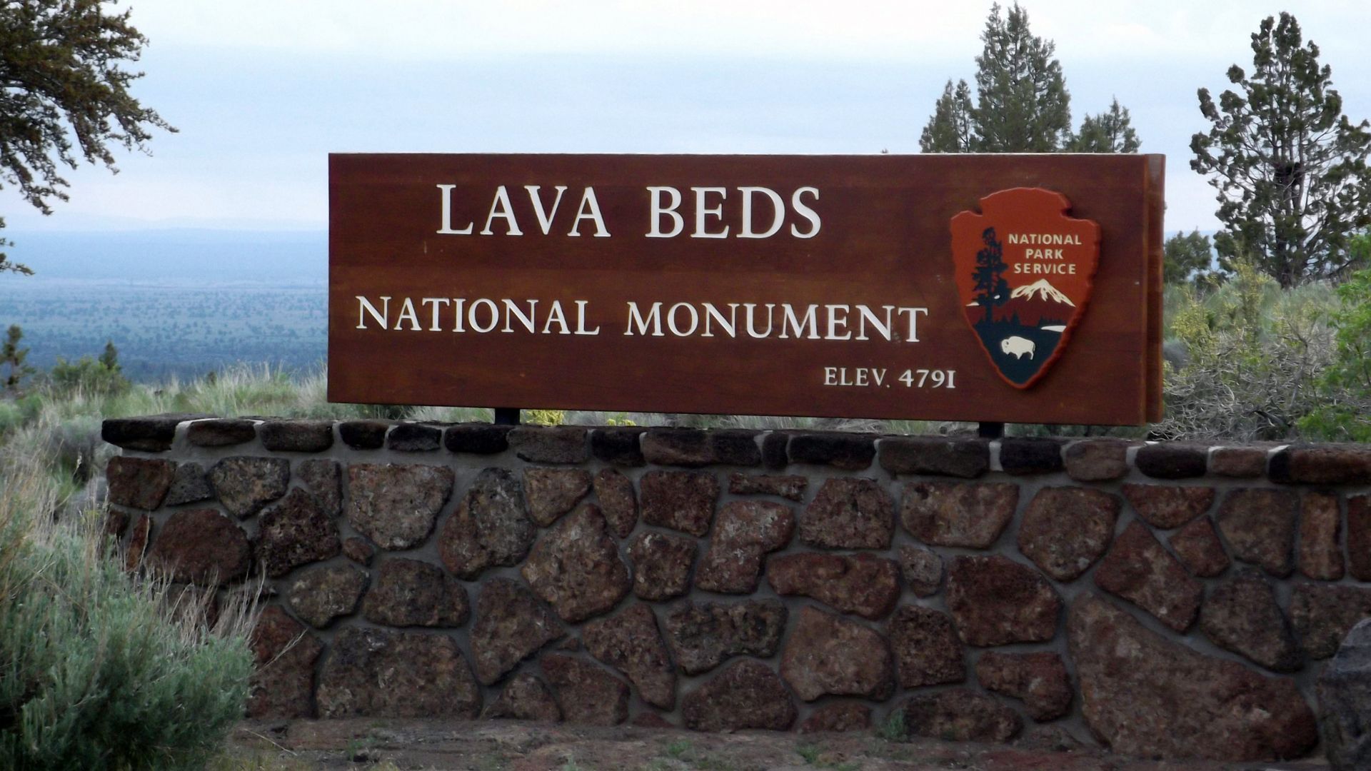 A wooden sign for Lava Beds National Monument stands on a stone wall, with the text "LAVA BEDS NATIONAL MONUMENT," "ELEV. 4791," and the National Park Service emblem visible against a hazy sky and sparse vegetation.