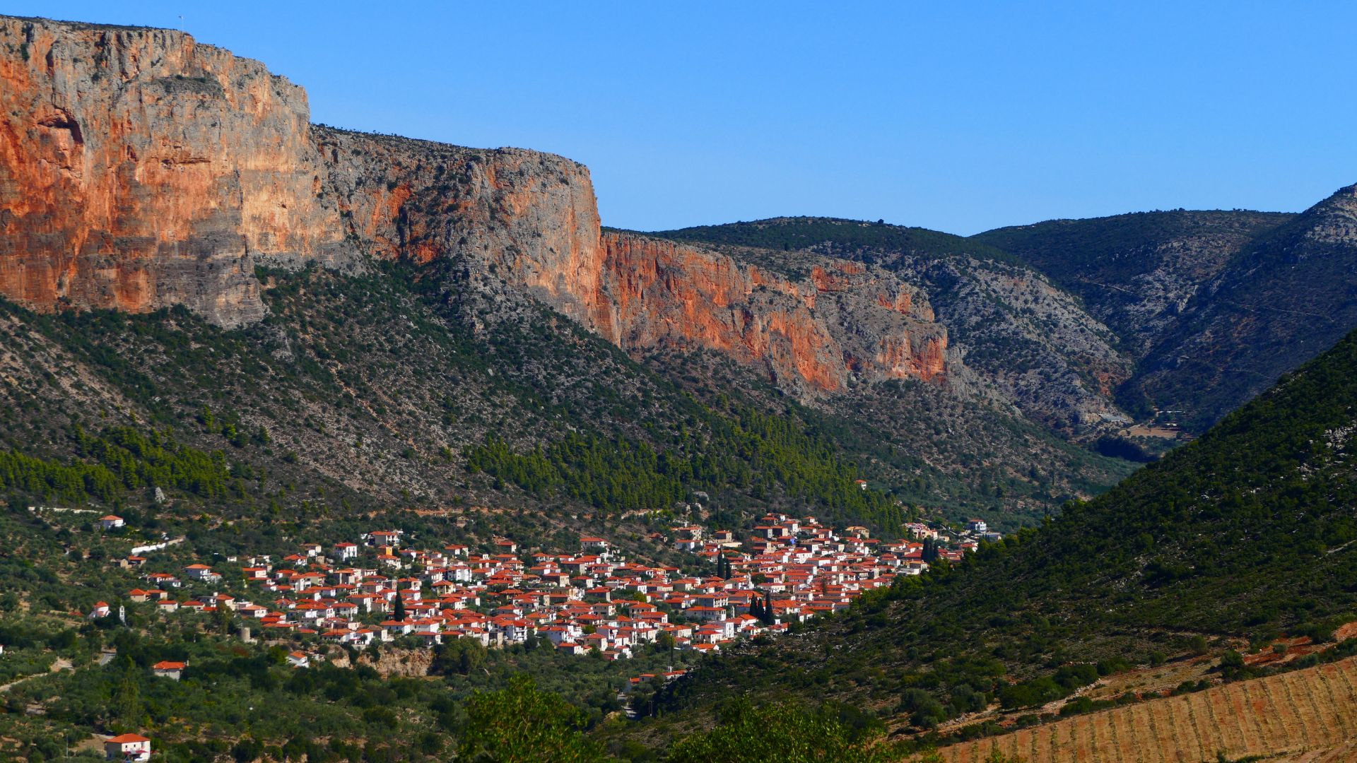 A small town with white buildings and red-tiled roofs nestled in a valley between imposing, reddish-brown cliffs and green, tree-covered mountains under a clear blue sky. 